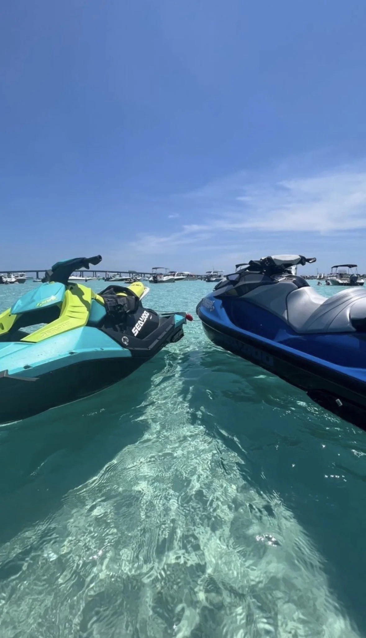 Two jet skis floating on clear turquoise water with a clear blue sky and boats in the background.