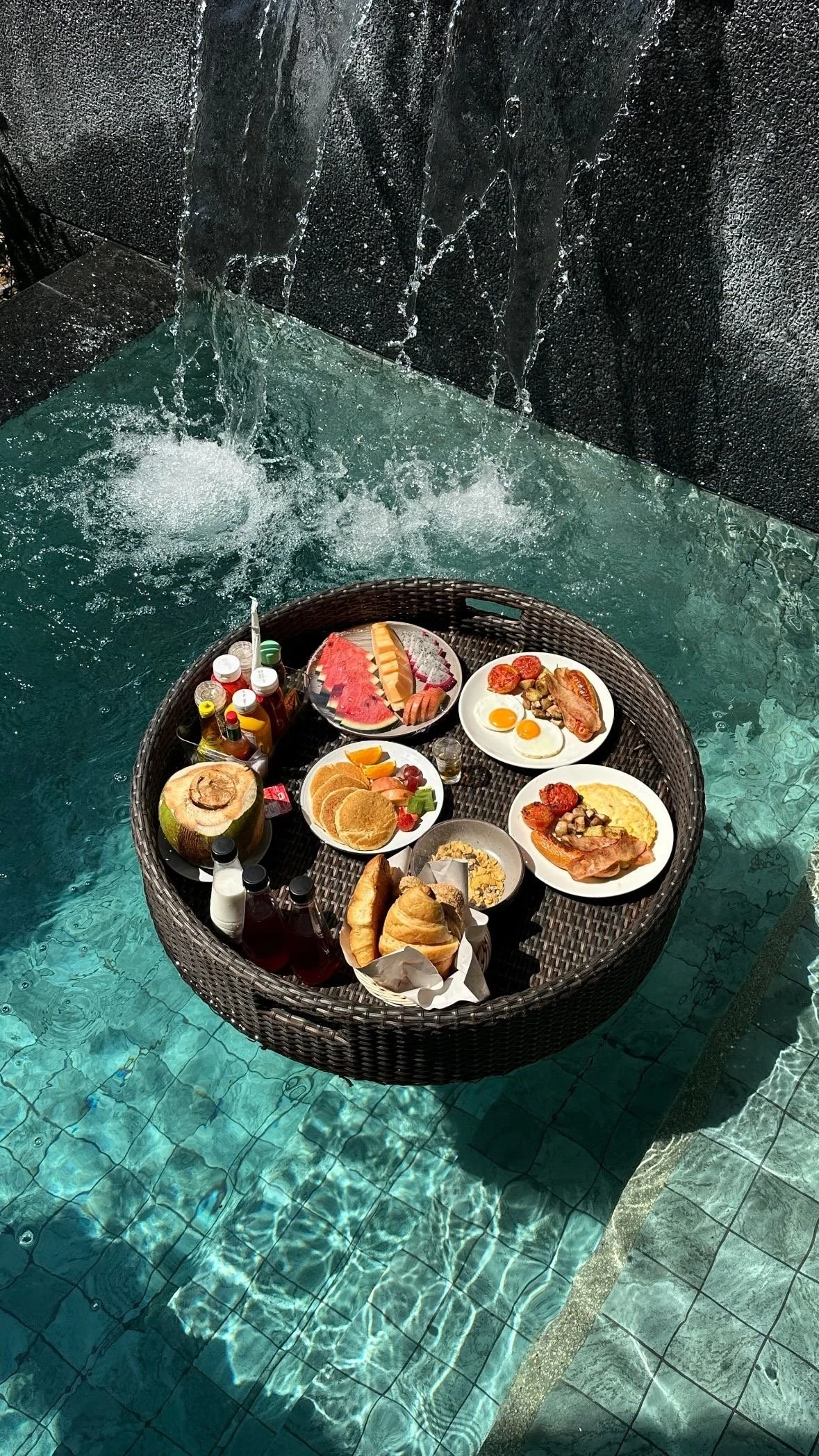 A floating wicker tray in a swimming pool holds breakfast items including various plates of eggs, bacon, sausage, watermelon, pancakes, croissants, bread rolls, juice, condiments, and fruit.