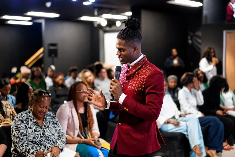 Prophet Jahhel Chosen in a red embellished blazer, ministering with a microphone among a seated congregation
