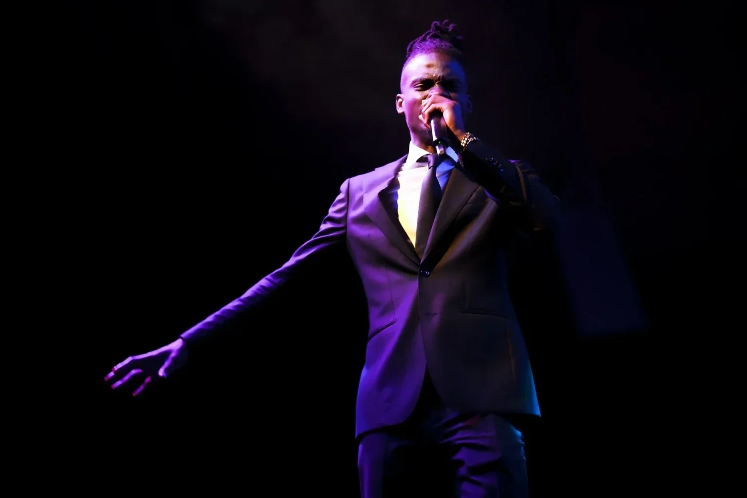 Prophet Jahhel Chosen ministering on a dark stage under violet light, microphone raised, one arm stretched toward the room