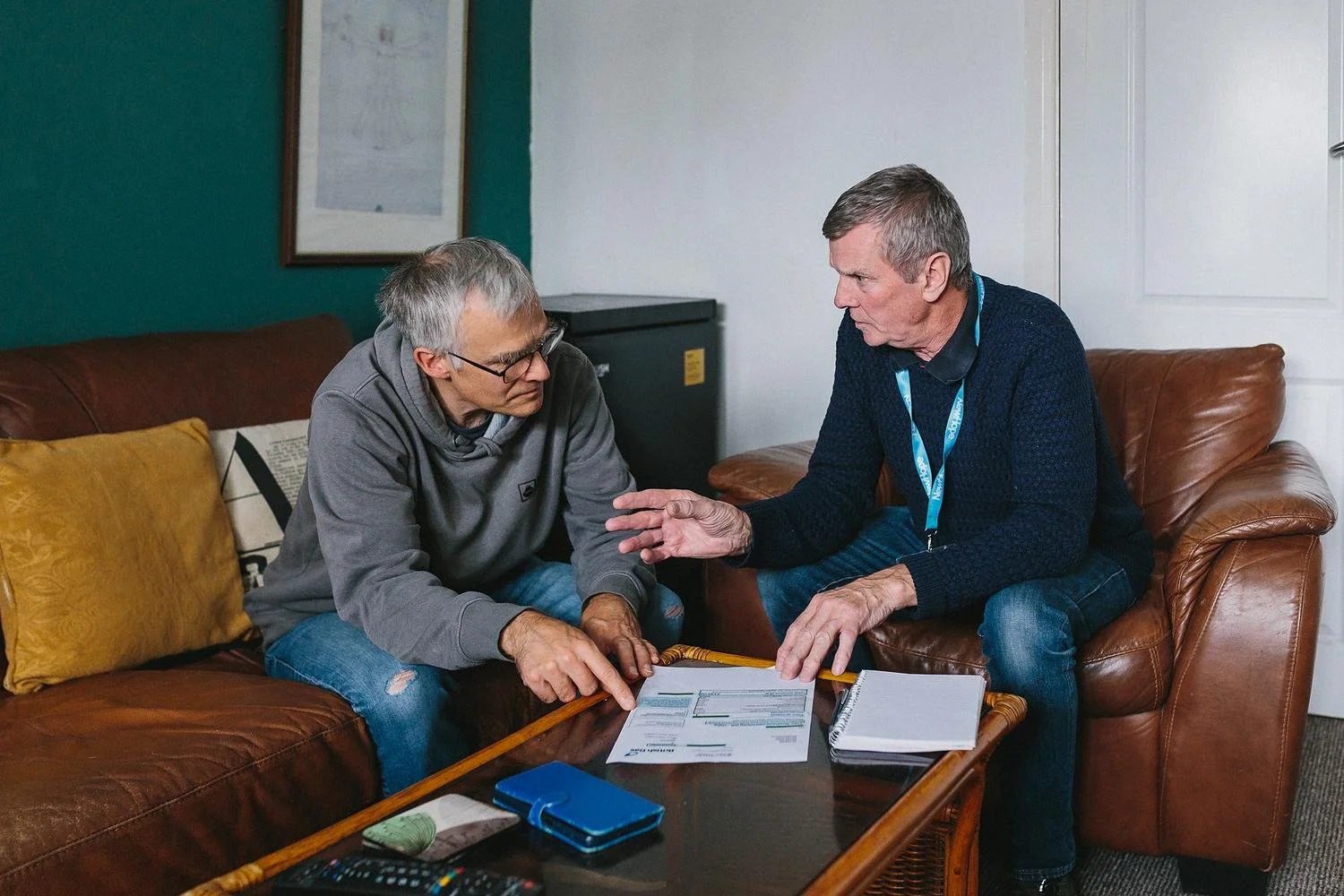 Two men sitting on a couch, one of them pointing at a piece of paper.
