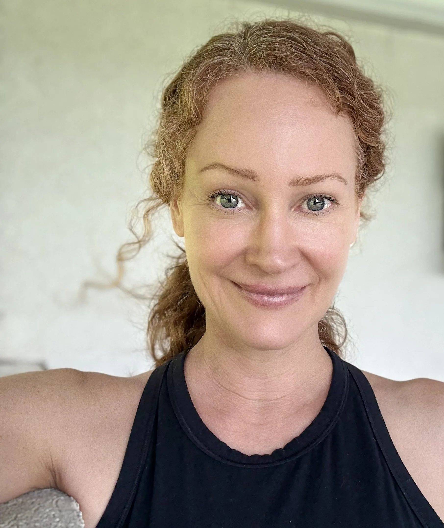 Close-up of a woman with curly red hair and green eyes, smiling while taking a selfie in a black athletic top.