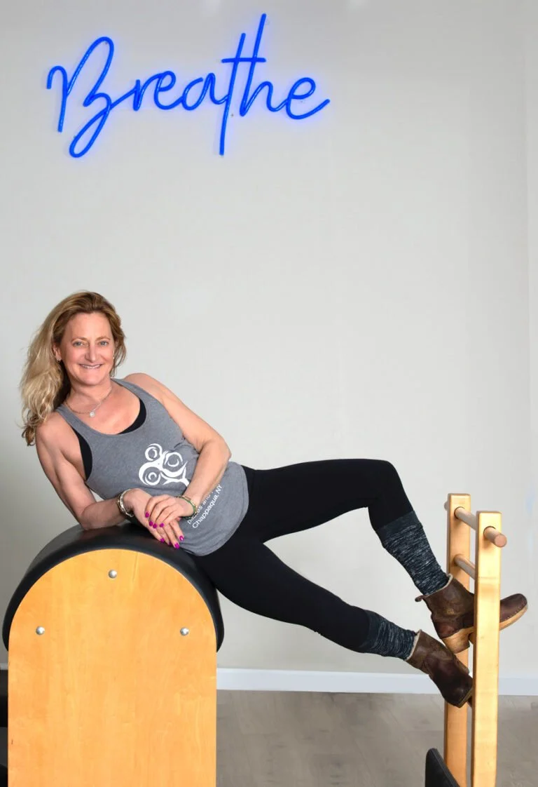 A woman in fitness attire lying on a Pilates barrel, with a large blue neon sign reading 'Breathe' above her. The background is a plain white wall.