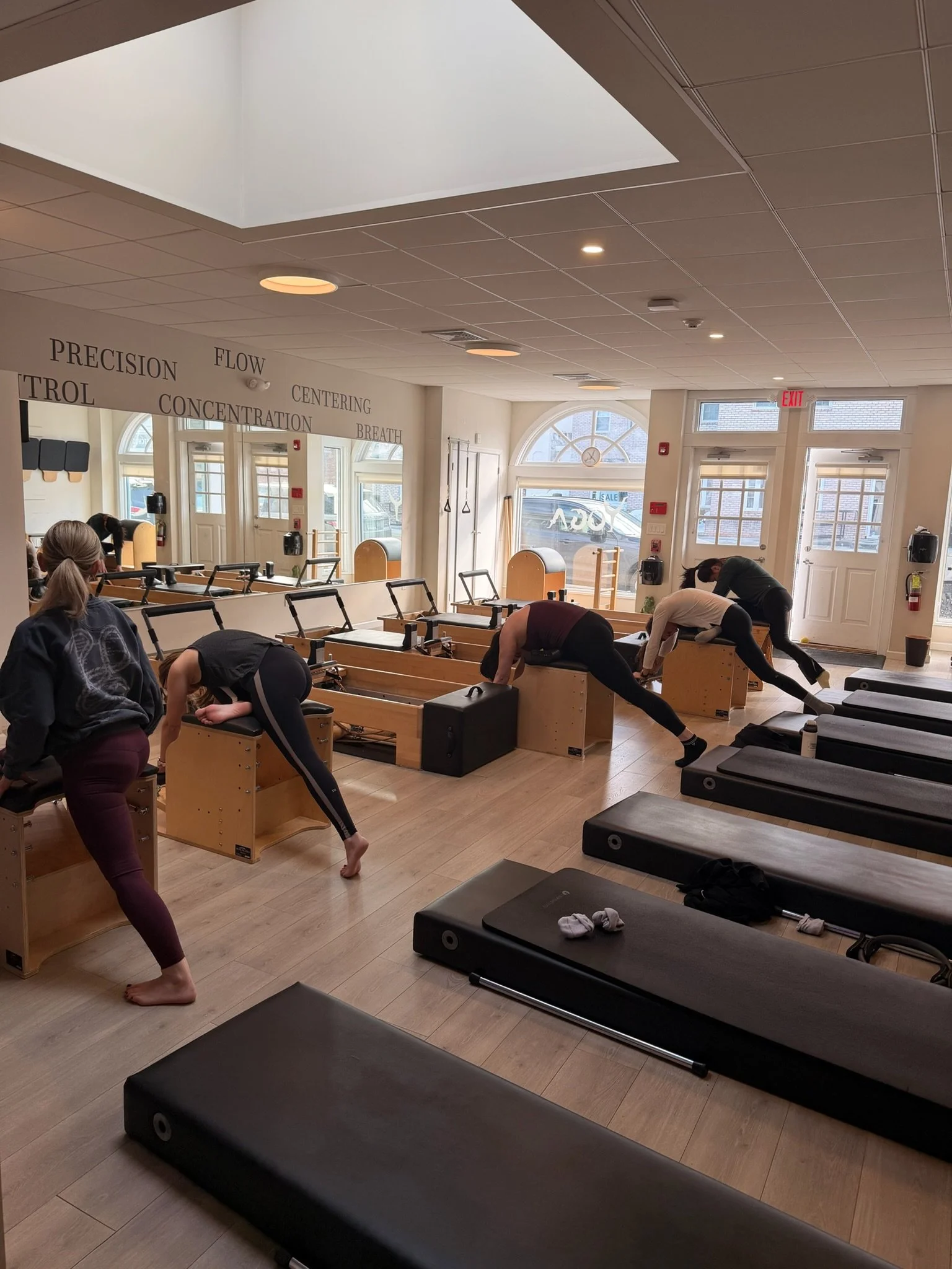 People participating in a Pilates class in a bright, spacious studio with large windows, wooden flooring, and wall decor with motivational words.