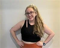 A young woman with long, wavy blonde hair wearing glasses, a black sleeveless crop top, and a black skirt, standing against a plain light-colored wall.