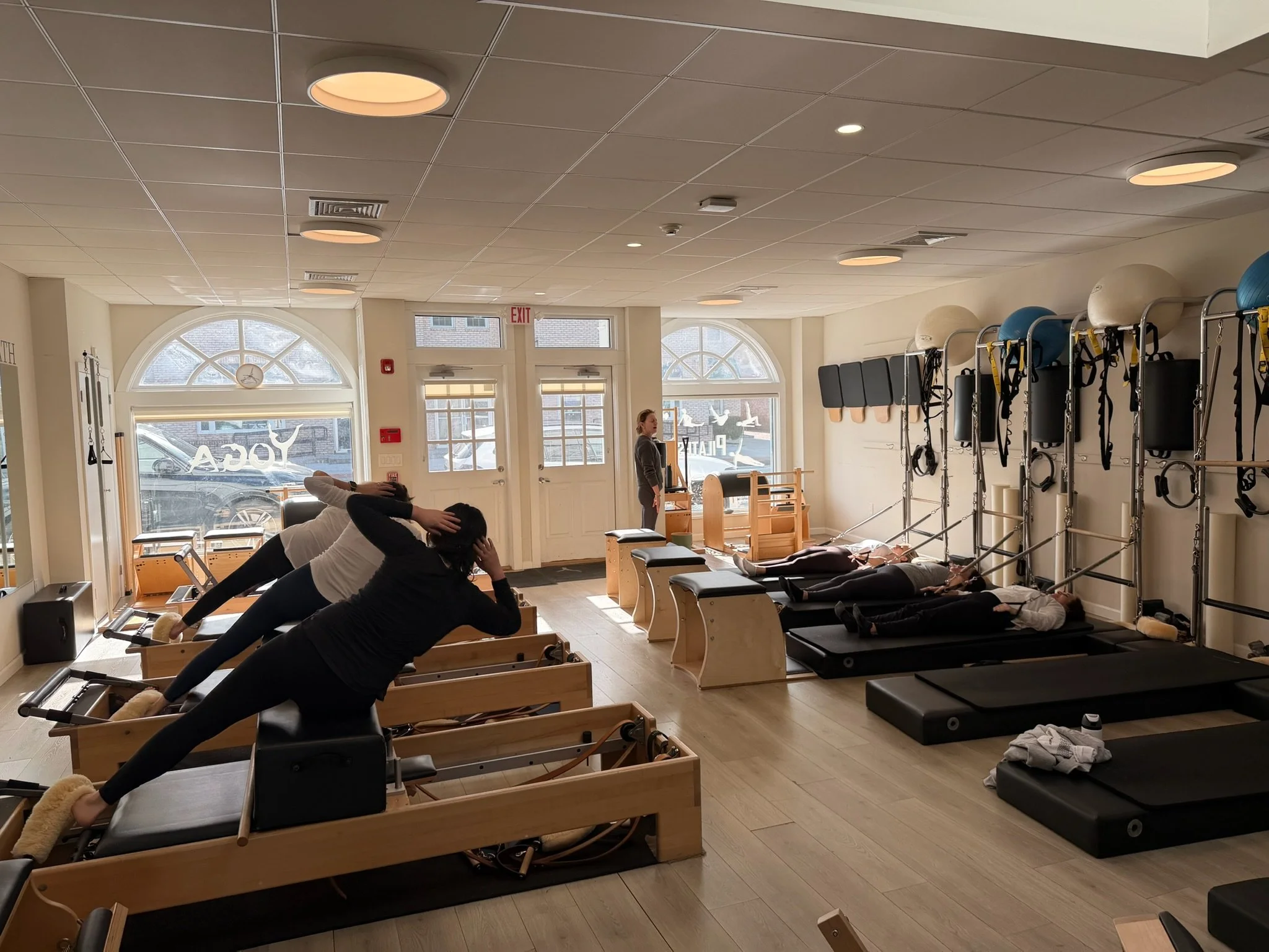 People exercising on Pilates reformer machines in a fitness studio with large windows, wall-mounted therapy balls, and fitness equipment.