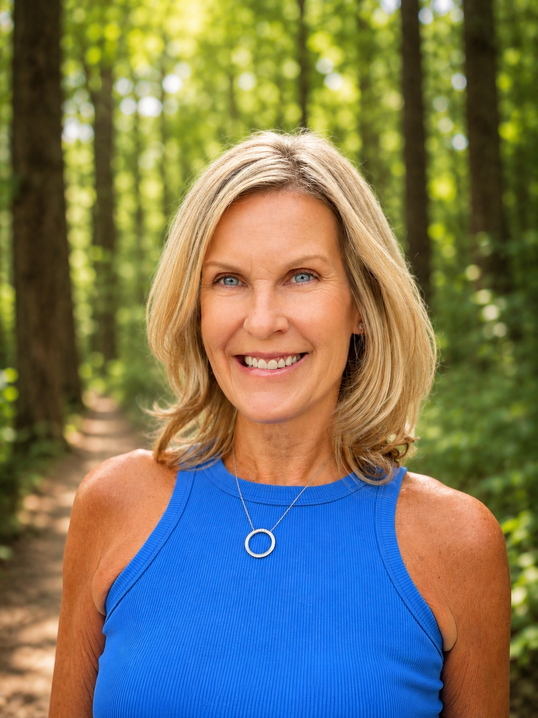 Smiling woman with blonde hair wearing a bright blue sleeveless top in a forest setting.