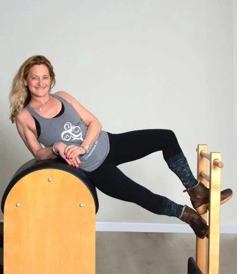 A woman with blonde hair lying on her side on Pilates equipment, smiling at the camera, wearing a gray tank top, black leggings, and brown ankle boots.