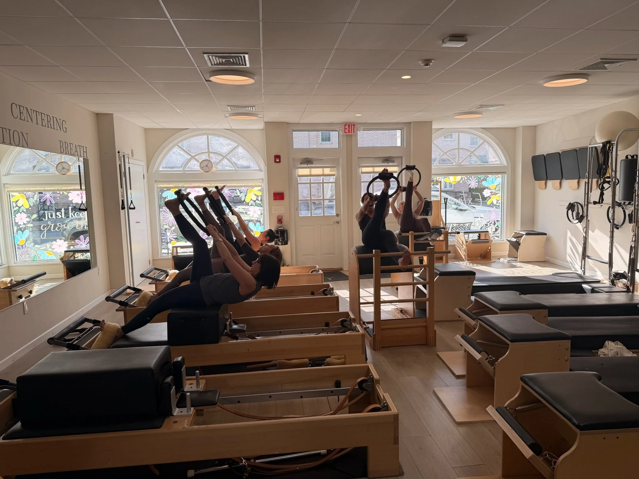 An interior view of a Pilates or fitness studio with participants using reformer machines for exercise, sunlight streaming through decorated windows, and motivational words on the wall.