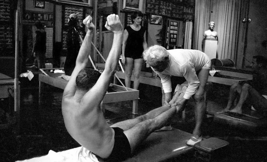 A man with muscular build doing a stretching exercise on the floor while an elderly woman assists him by holding his legs in a gym or dance studio. Other people are visible in the background, some stretching and some standing near gym equipment.