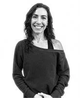 A woman with shoulder-length curly hair smiling, dressed in a black off-the-shoulder top, standing against a plain background.