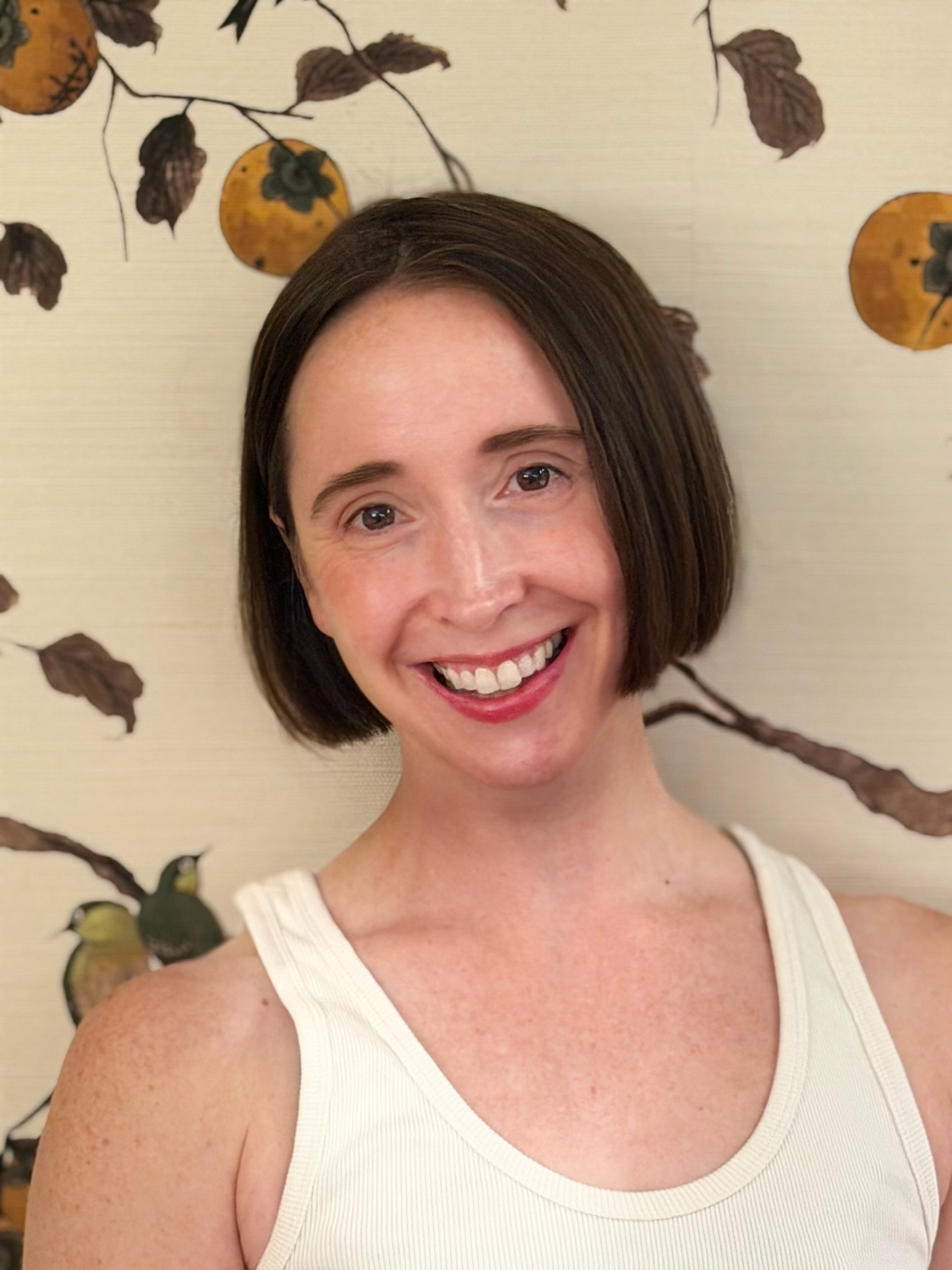 Close-up of a smiling woman with short brown hair, wearing a white sleeveless top, against a background with a floral and fruit pattern.