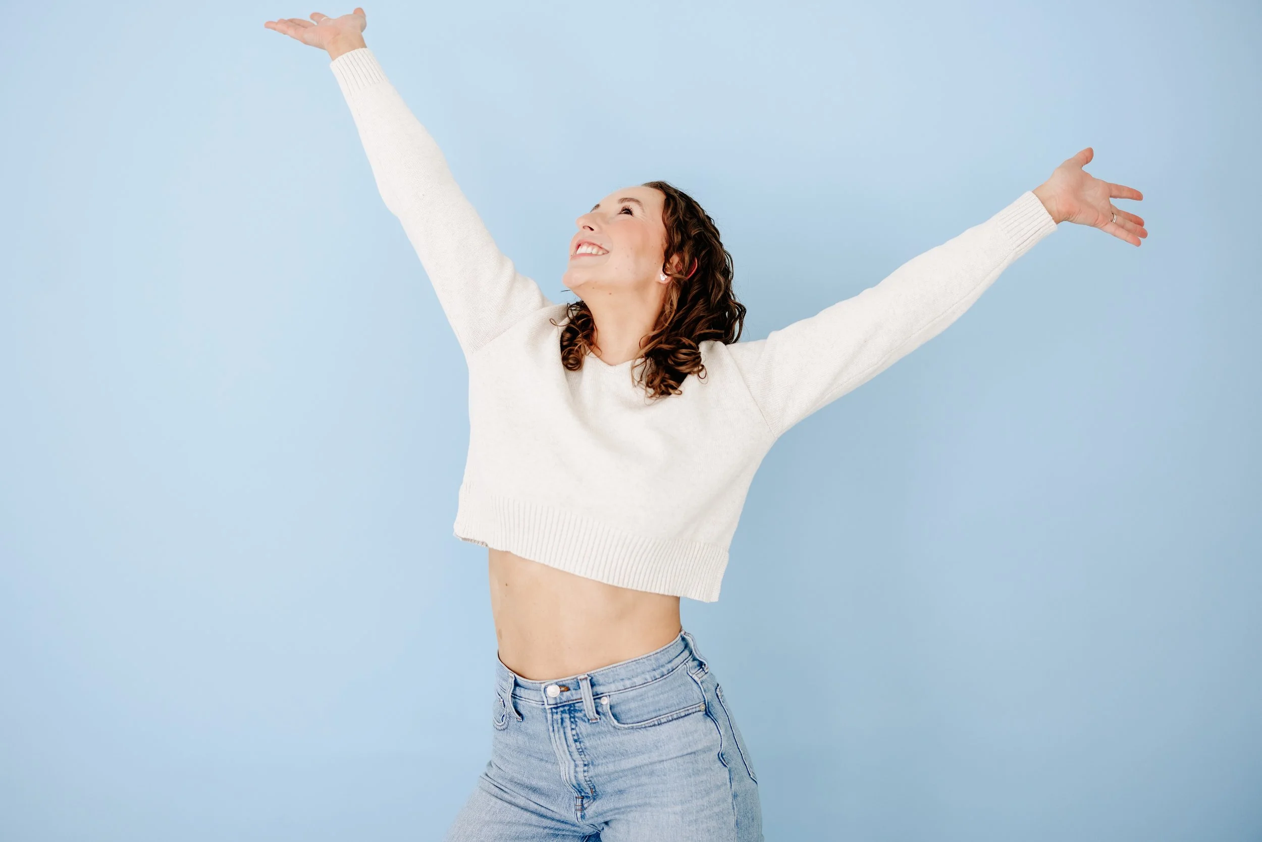 Curly-haired woman in a cream sweater and jeans smiling with arms outstretched against a blue background.