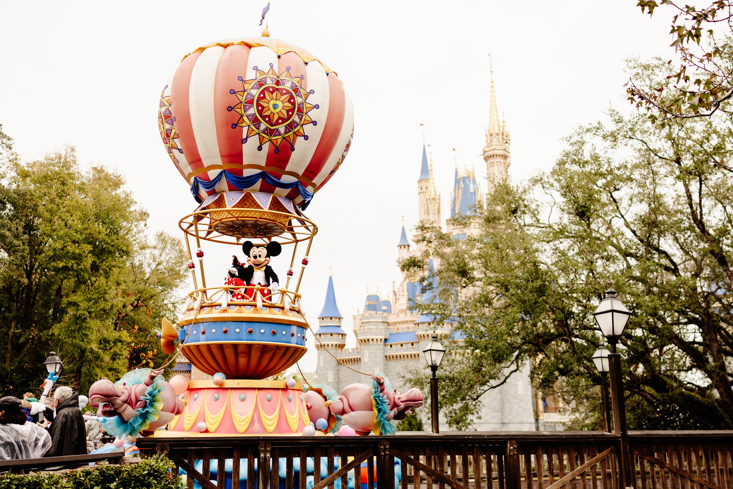 Mickey and Minnie waving from a hot air balloon parade float with Cinderella Castle in the background.