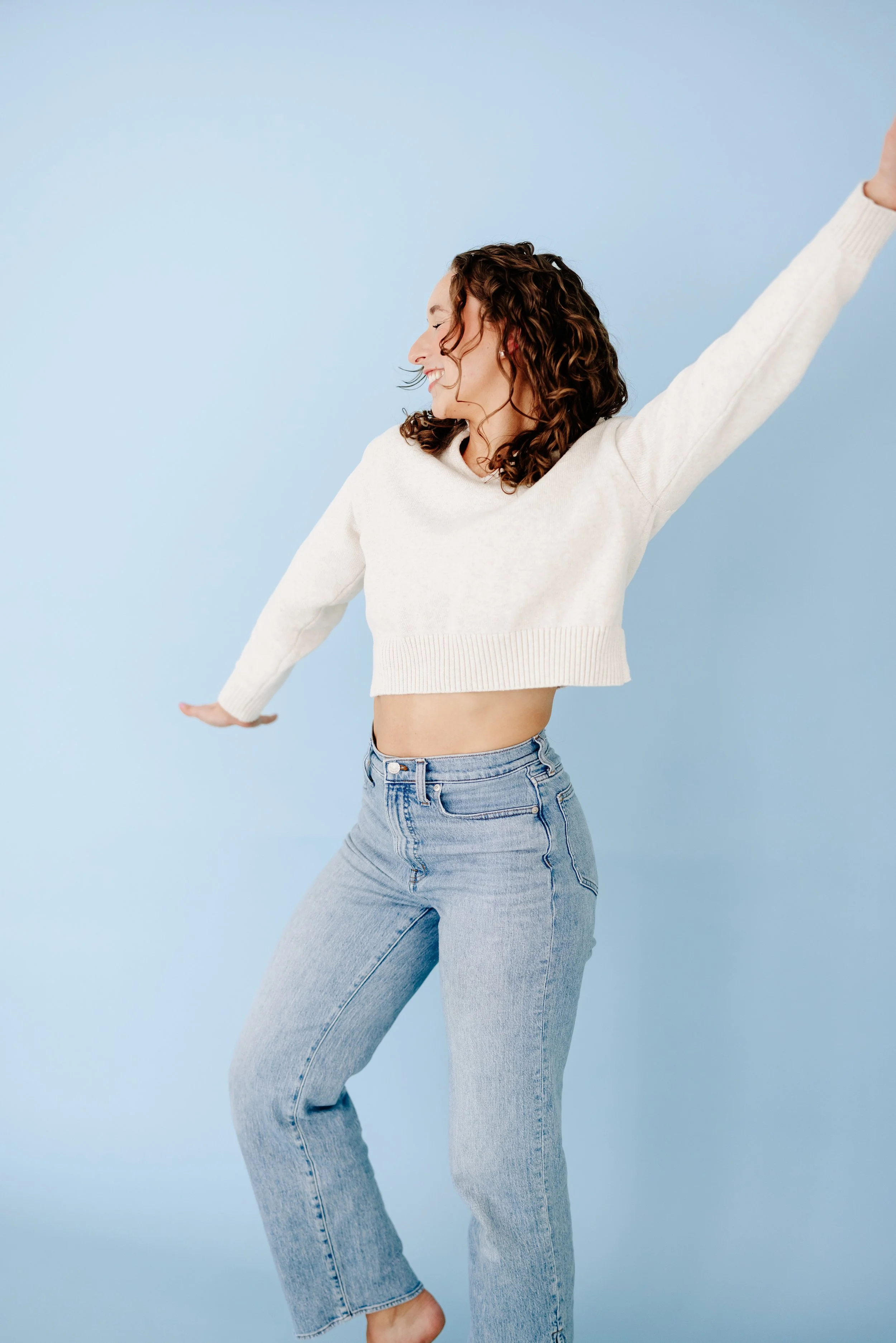 Curly-haired woman in a cream sweater and jeans smiling with arms outstretched against a blue background.