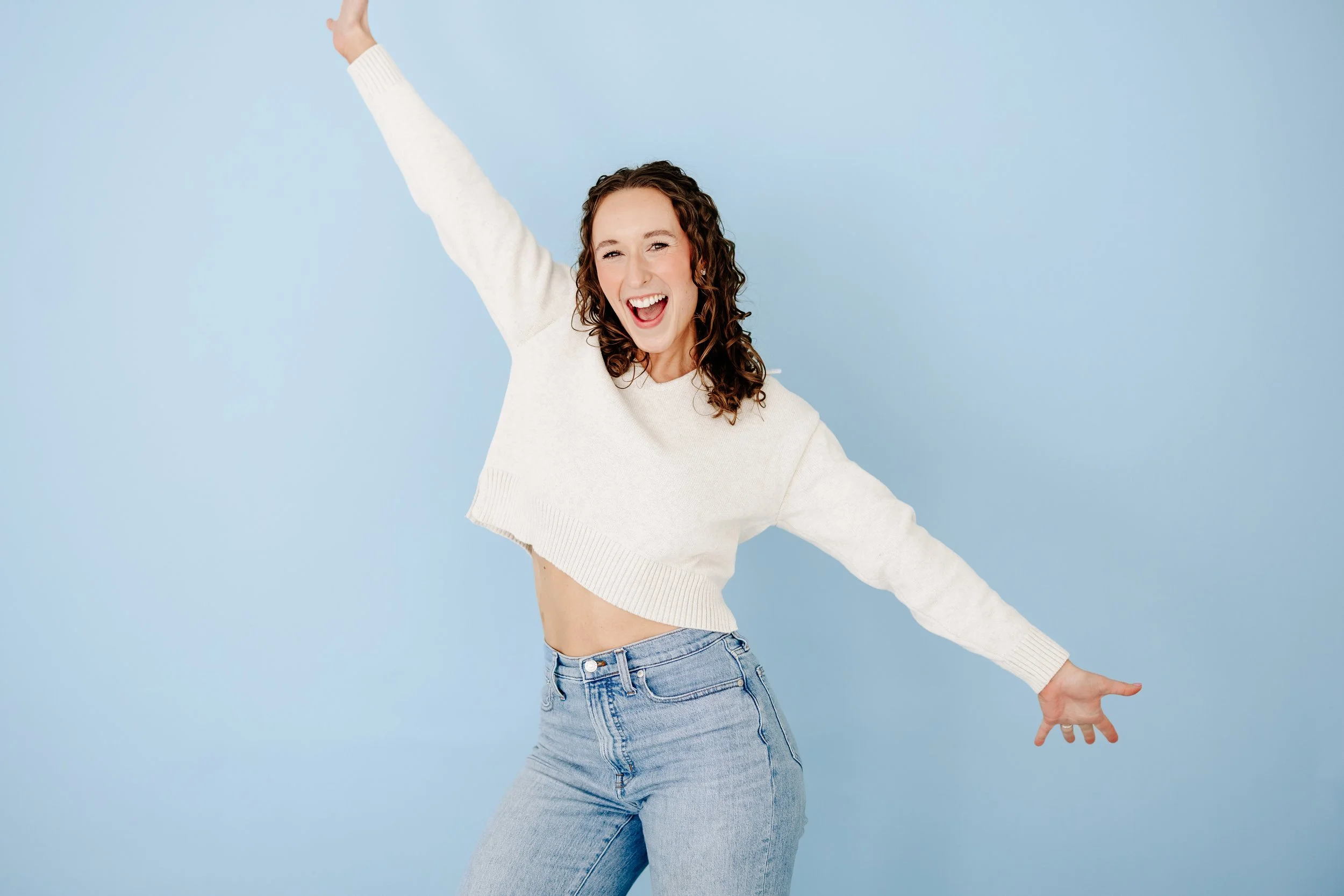 Curly-haired woman in a cream sweater and jeans smiling with arms outstretched against a blue background.