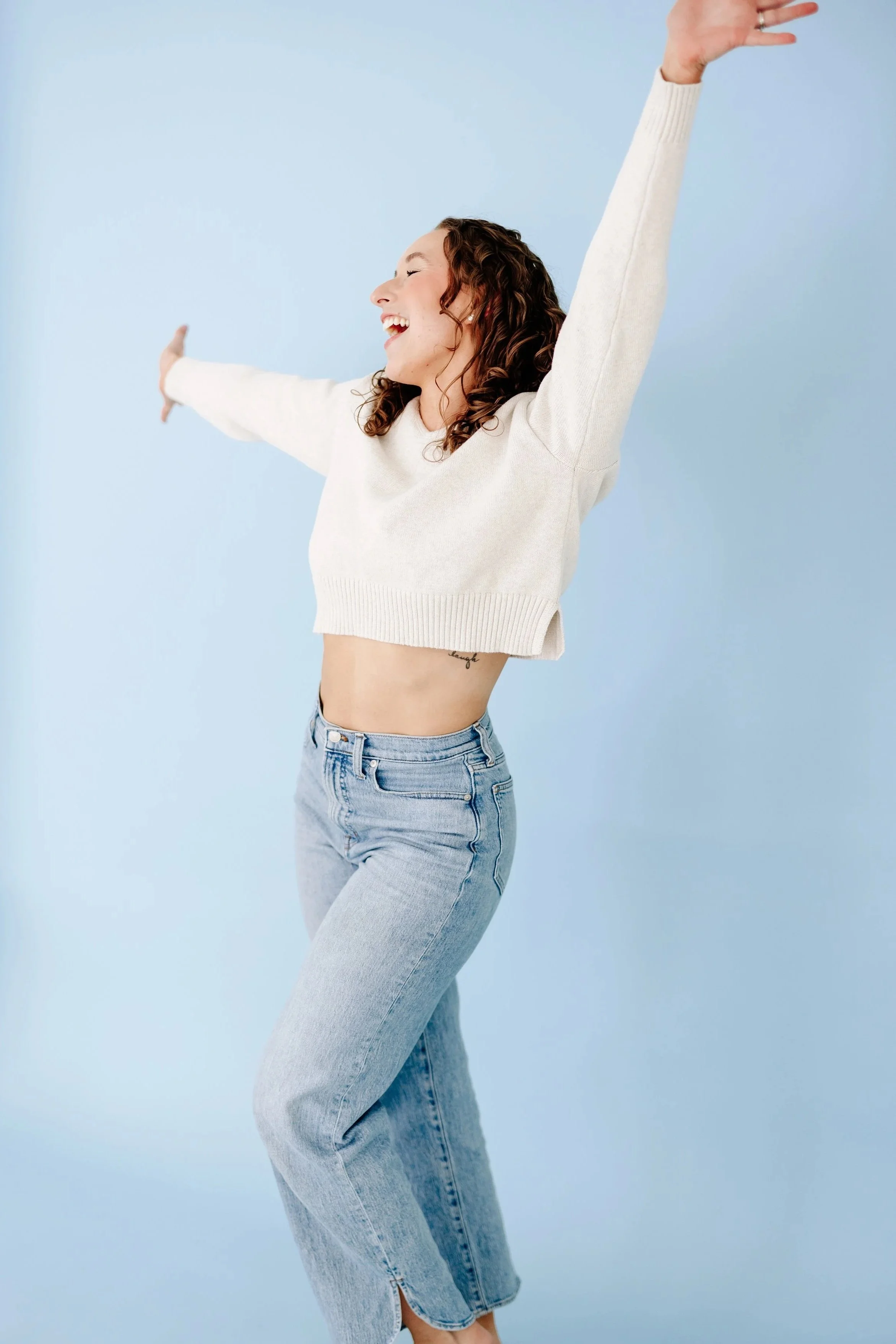 Curly-haired woman in a cream sweater and jeans smiling with arms outstretched against a blue background.