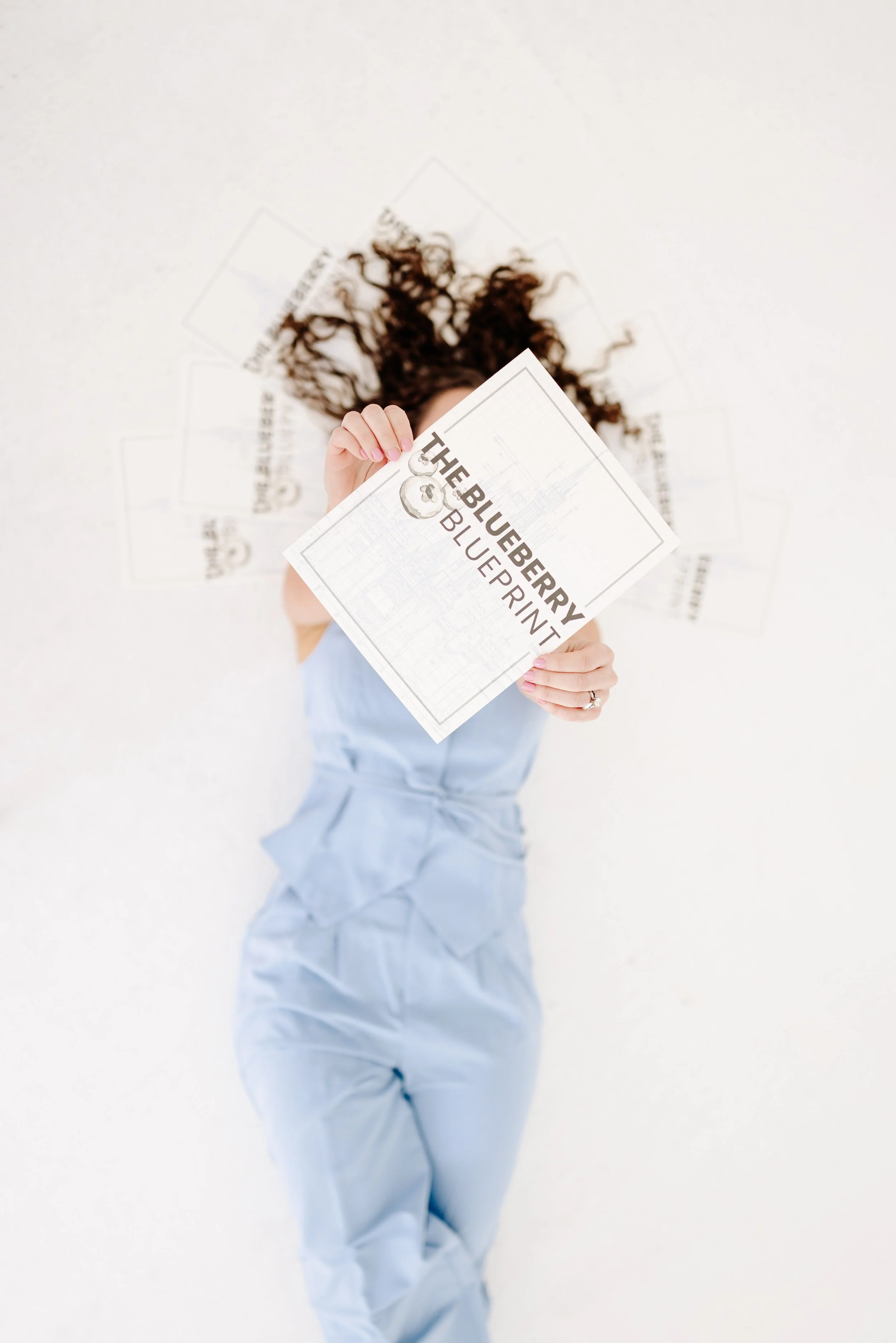 Woman lying down holding up The Blueberry Blueprint course workbook, surrounded by printed copies fanned around her