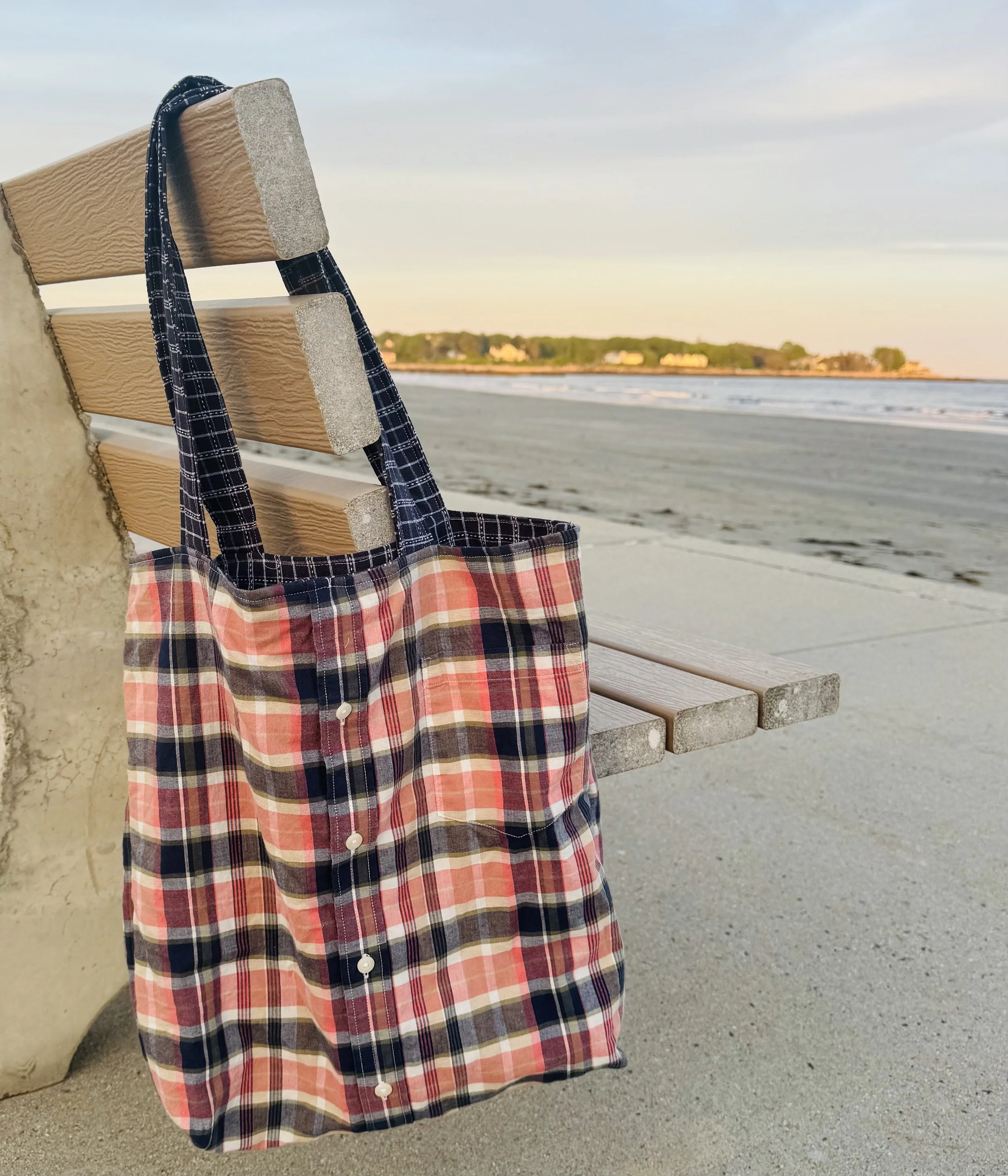 A plaid bag hanging on a wooden bench at the beach with the ocean and distant trees in the background during sunset.