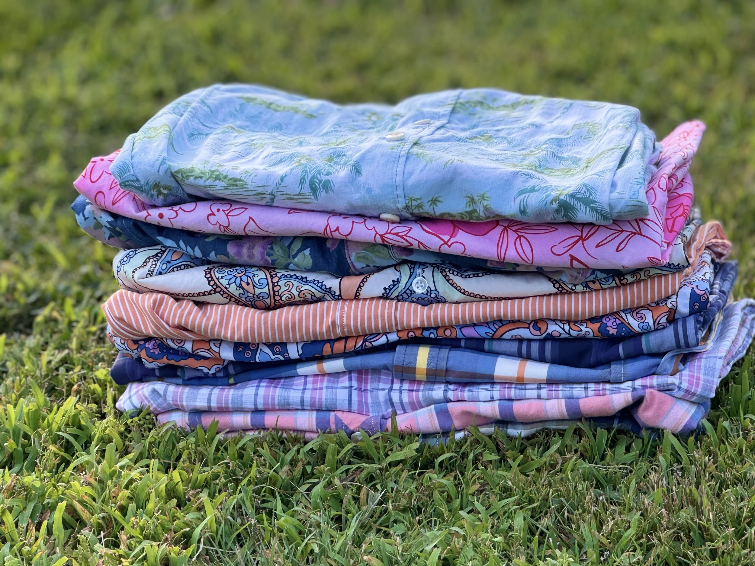 A stack of colorful, patterned shirts resting on grass in an outdoor setting.