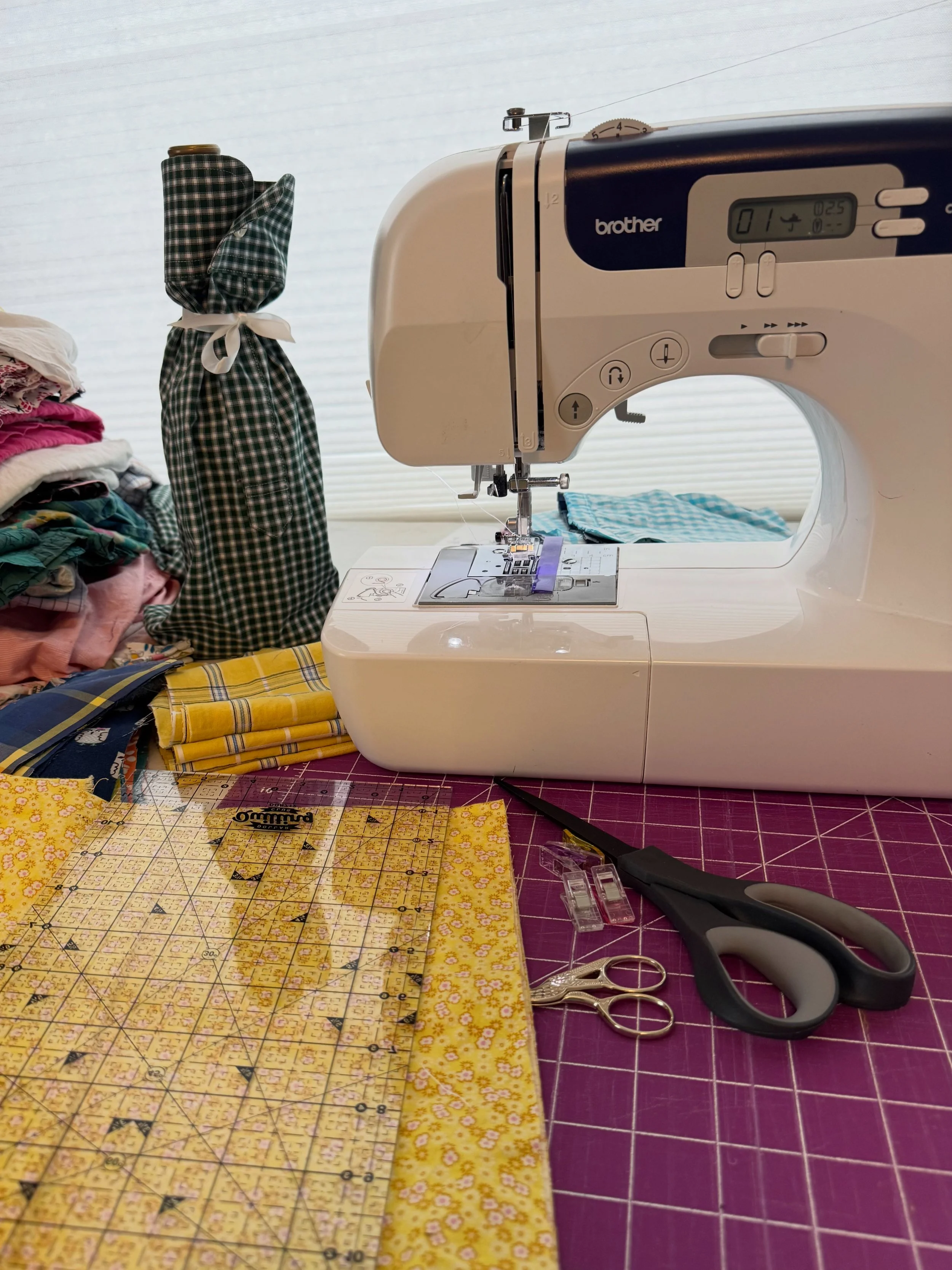 A sewing machine on a table with fabric and sewing supplies around, including scissors, scissors, a clear ruler, a yellow patterned fabric, and a pair of scissors. There is a stack of folded fabric on the left side.
