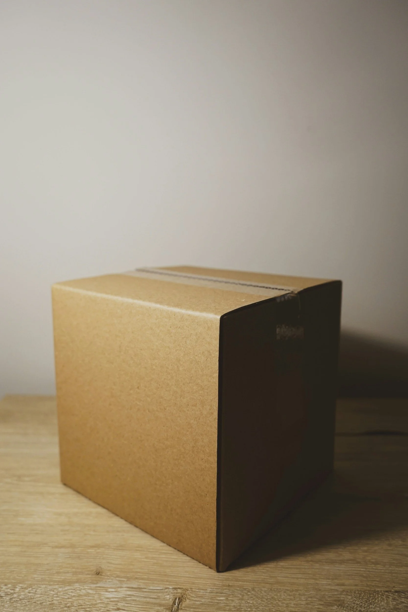 A plain cardboard box with a black object or packaging covering part of it, placed on a wooden surface with a light-colored background.