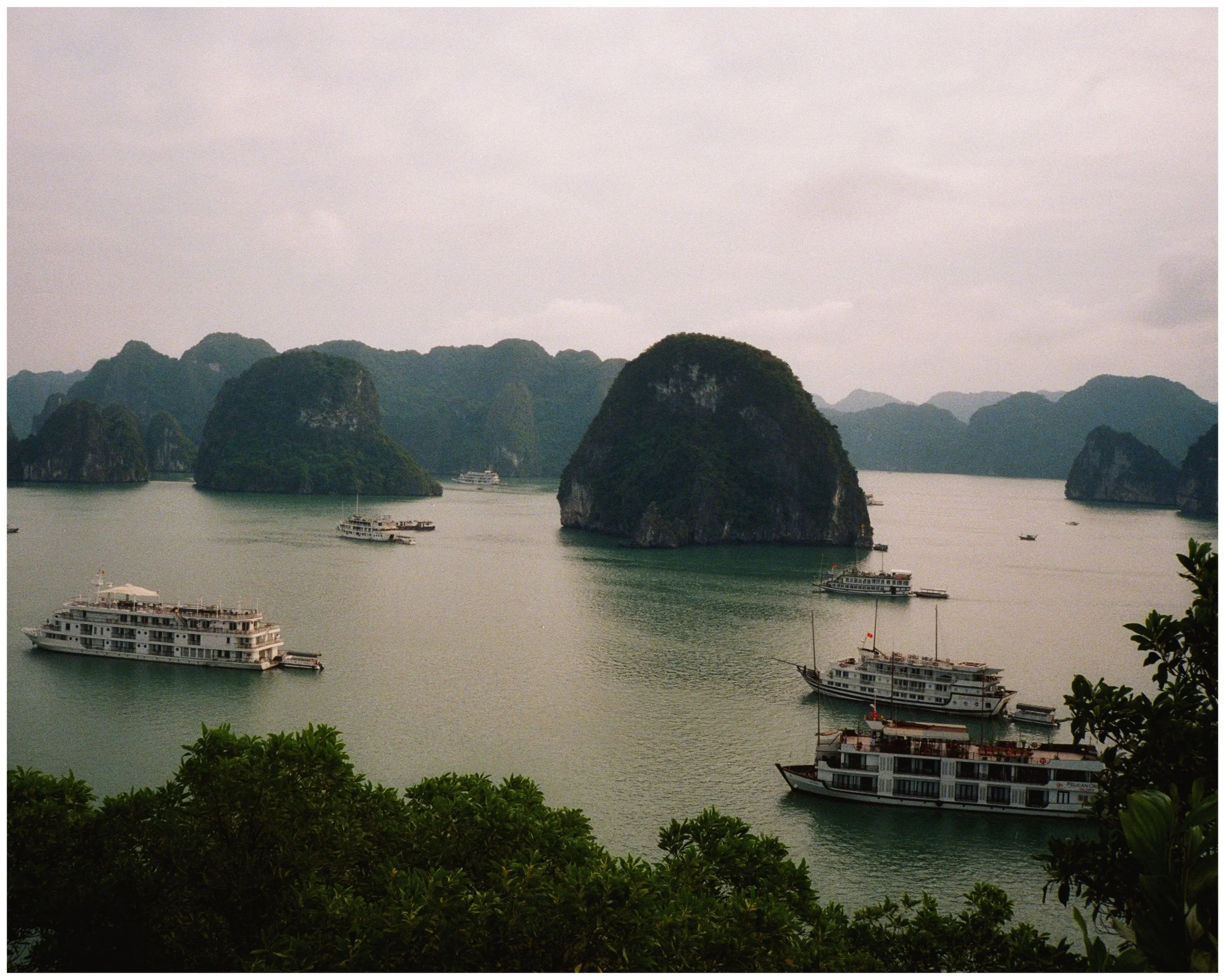 Several boats are sailing in a bay surrounded by tall, green, rocky islands with lush vegetation. The sky is overcast, and the water is calm.