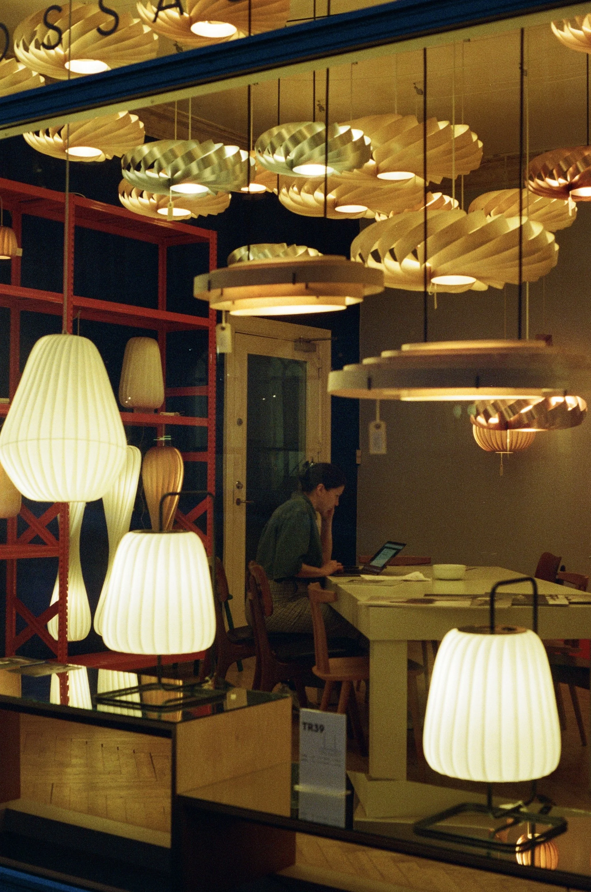 Interior lighting with modern pendant lamps hanging from the ceiling, some with warm yellow glow, in a restaurant or cafe setting. A woman is seated at a table, looking at a device or menu.