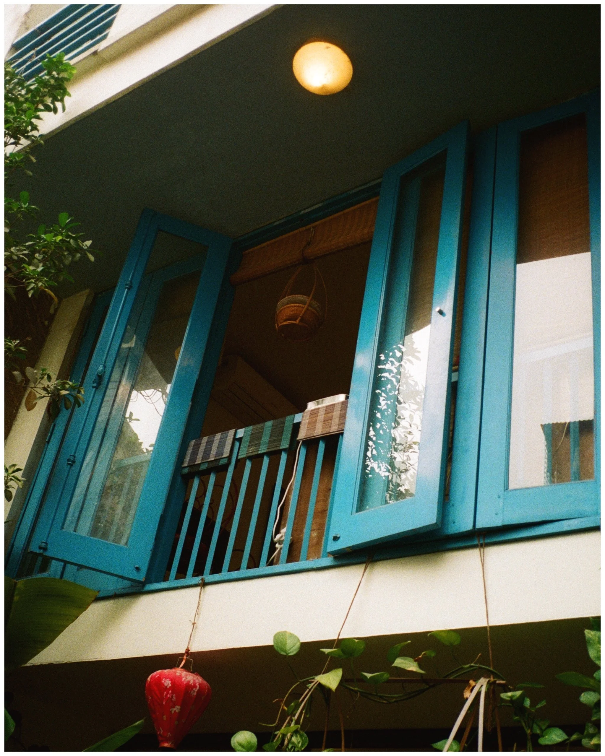 The photo shows a teal-colored balcony door with glass panels, open to the outside. There is a hanging woven basket on the balcony ceiling and a plaid curtain inside. A ceiling light is on above the balcony, and plants are visible in the foreground.