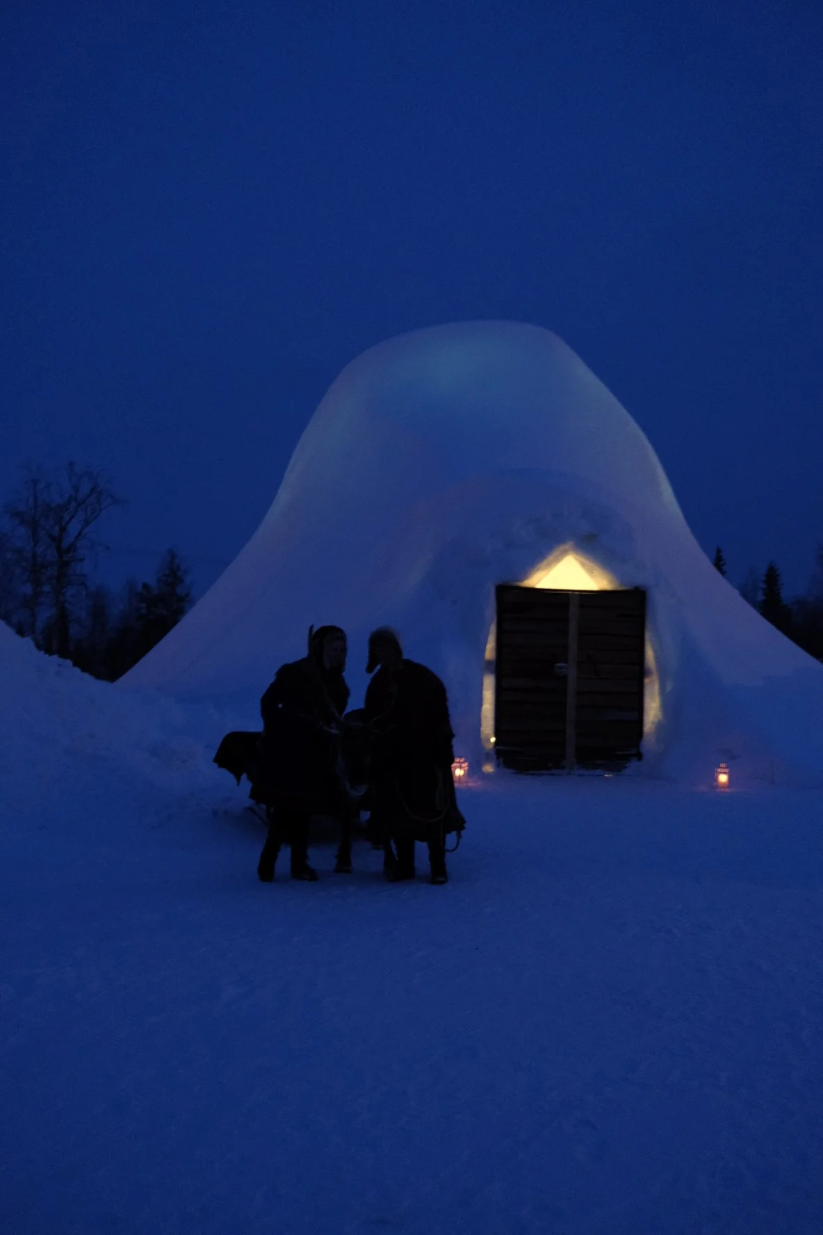 An igloo illuminated from within at dusk with two people standing outside, surrounded by snow and trees.