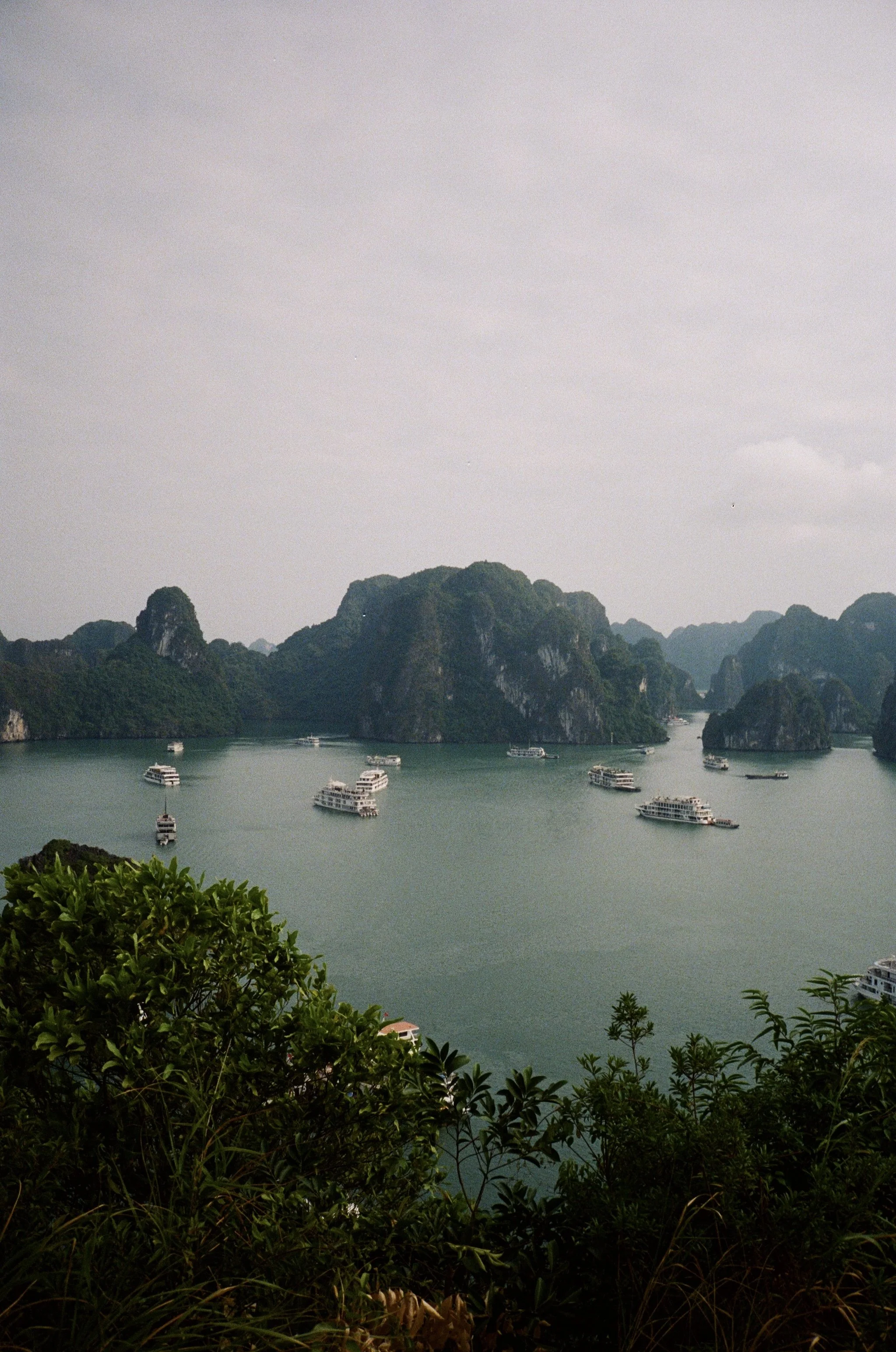 Multiple boats floating on a body of water surrounded by green mountains and lush vegetation, with an overcast sky.