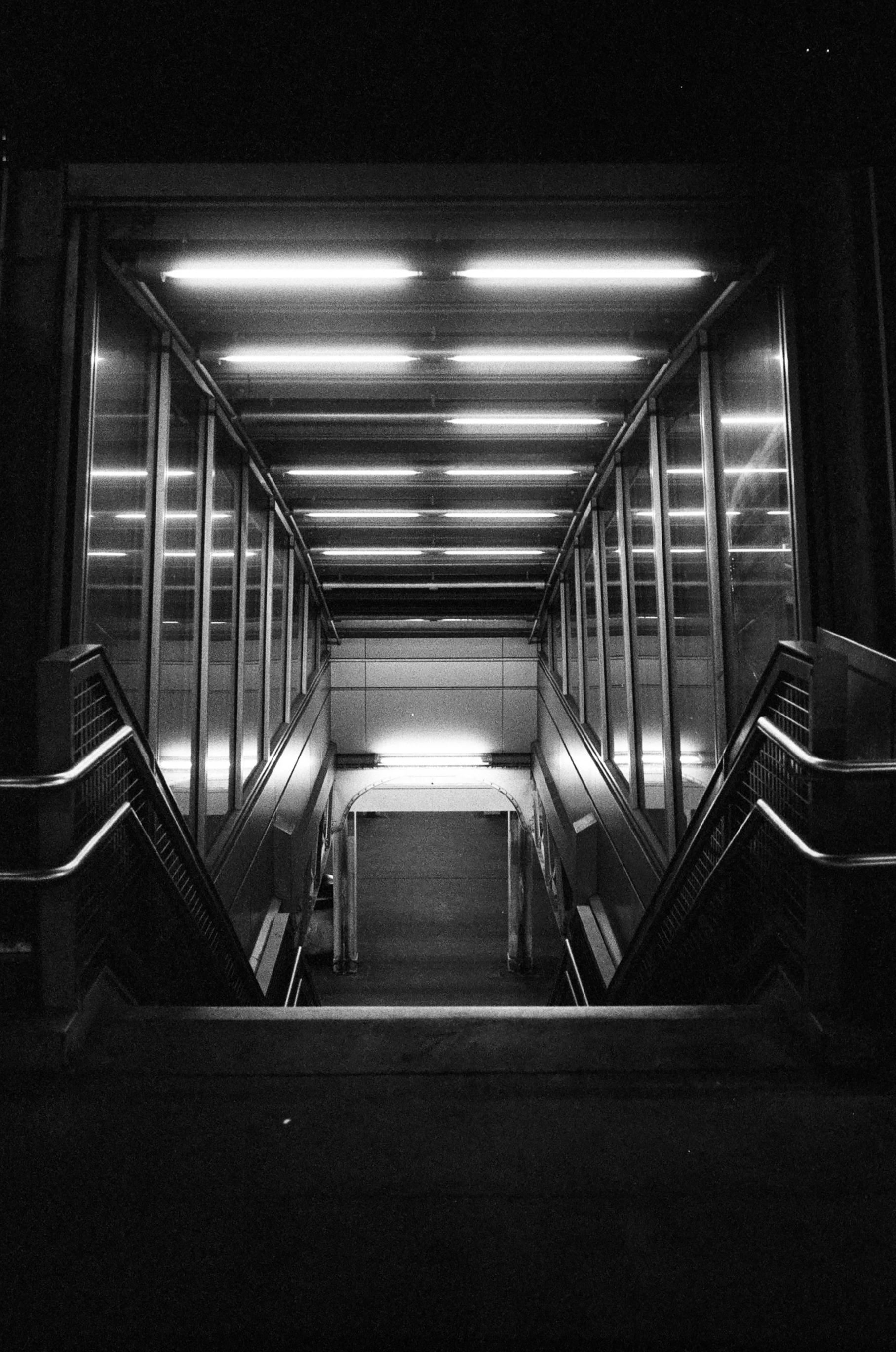 Black and white photo of an underground passageway or escalator area, illuminated by fluorescent lights on the ceiling, with metal railings on both sides and glass panels along the walls.