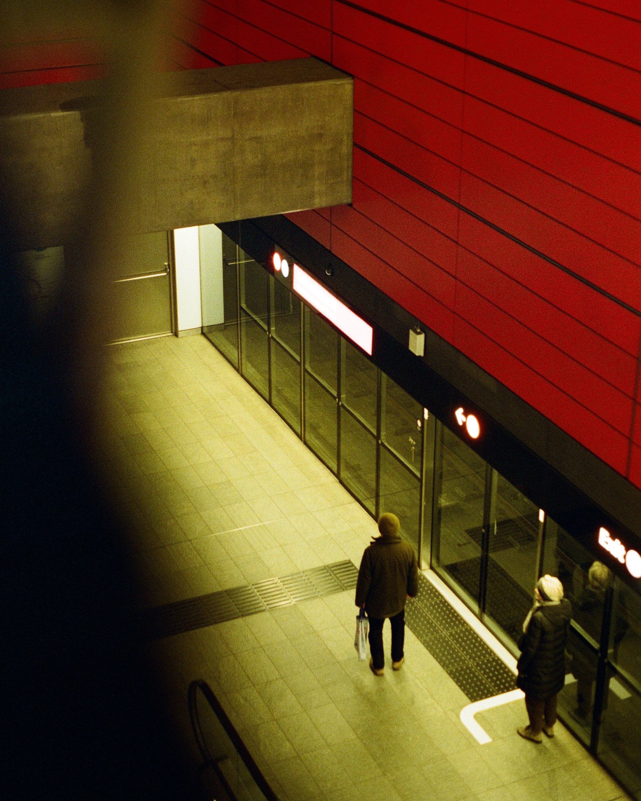 Two people walking into building with glass doors, red and black signage, and yellow floor tiles, at night or in low light.