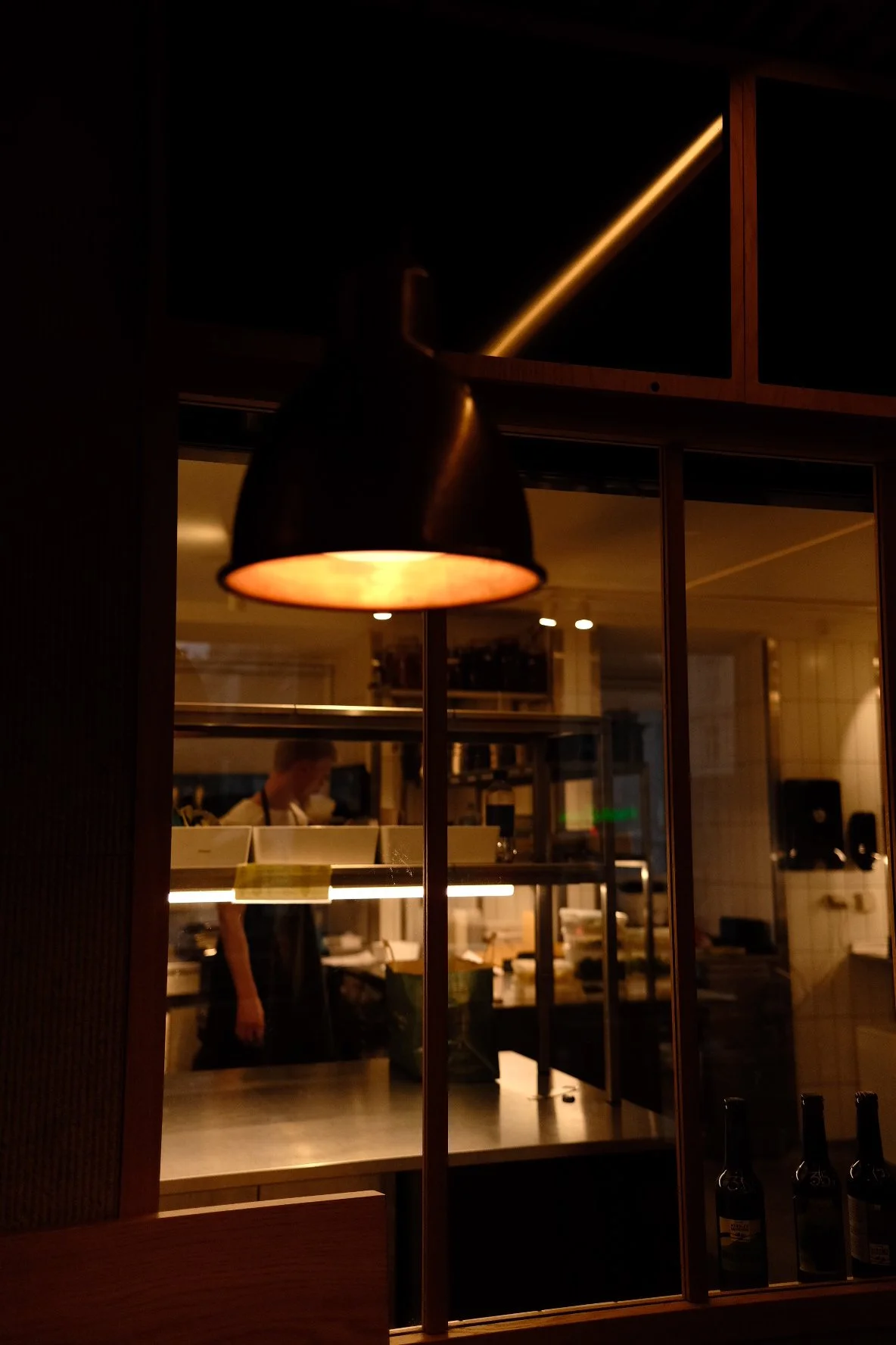 Indoor restaurant scene with a black hanging lamp casting warm light, a person working in the kitchen behind a glass window, and bottles on a table in the foreground.