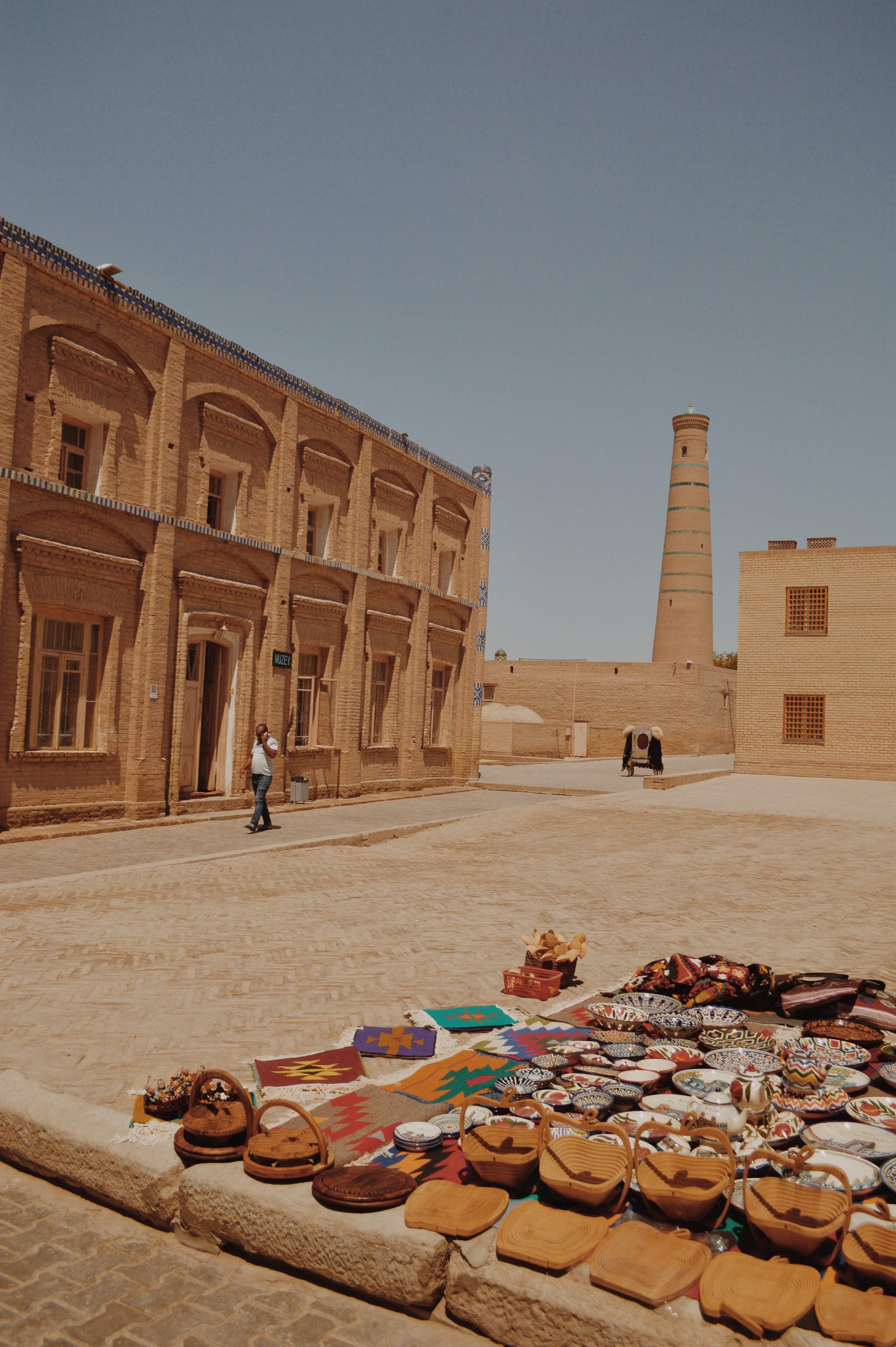 Market stall with colorful handmade crafts, pottery, and textiles displayed on a stone platform in an outdoor setting with historical architecture and a tall chimney in the background.