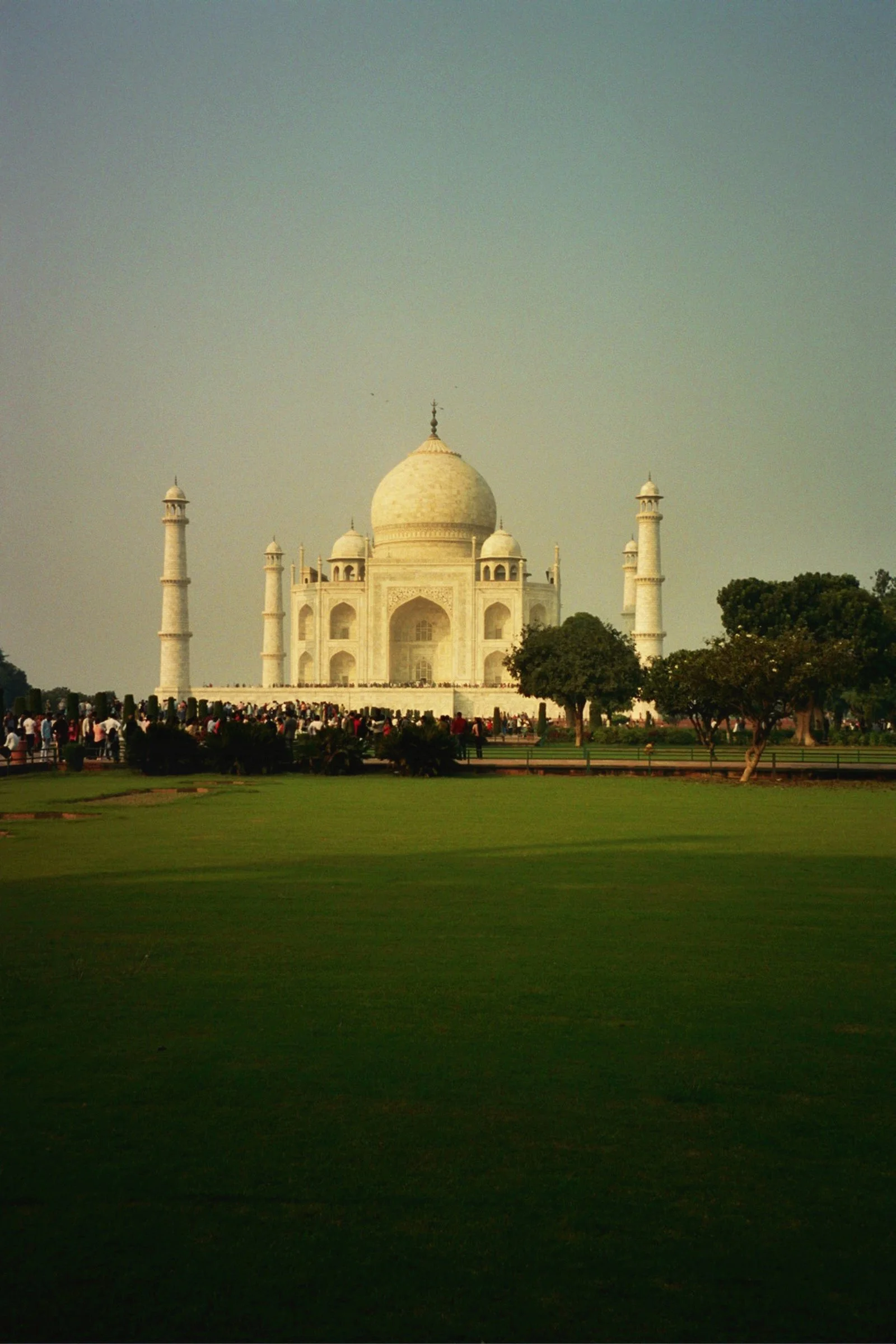 The Taj Mahal, a white marble mausoleum with a large central dome and four minarets, situated on a green lawn with trees and a crowd of visitors.
