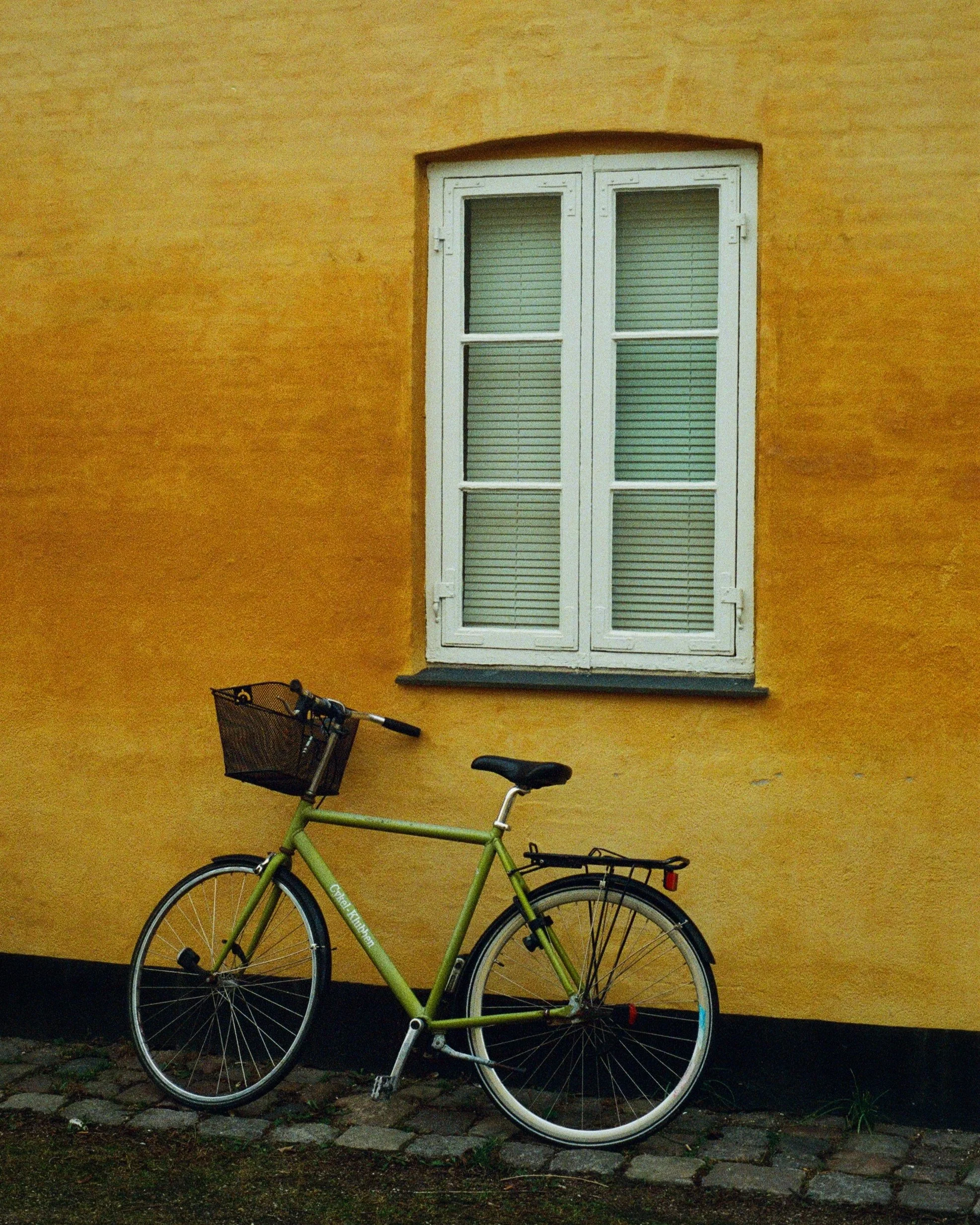 A lime green bicycle with a black basket attached to the front is parked against a yellow stucco wall. A white window with closed blinds is on the wall above the bicycle, and a small black step is below the window. The ground is paved with cobbleston