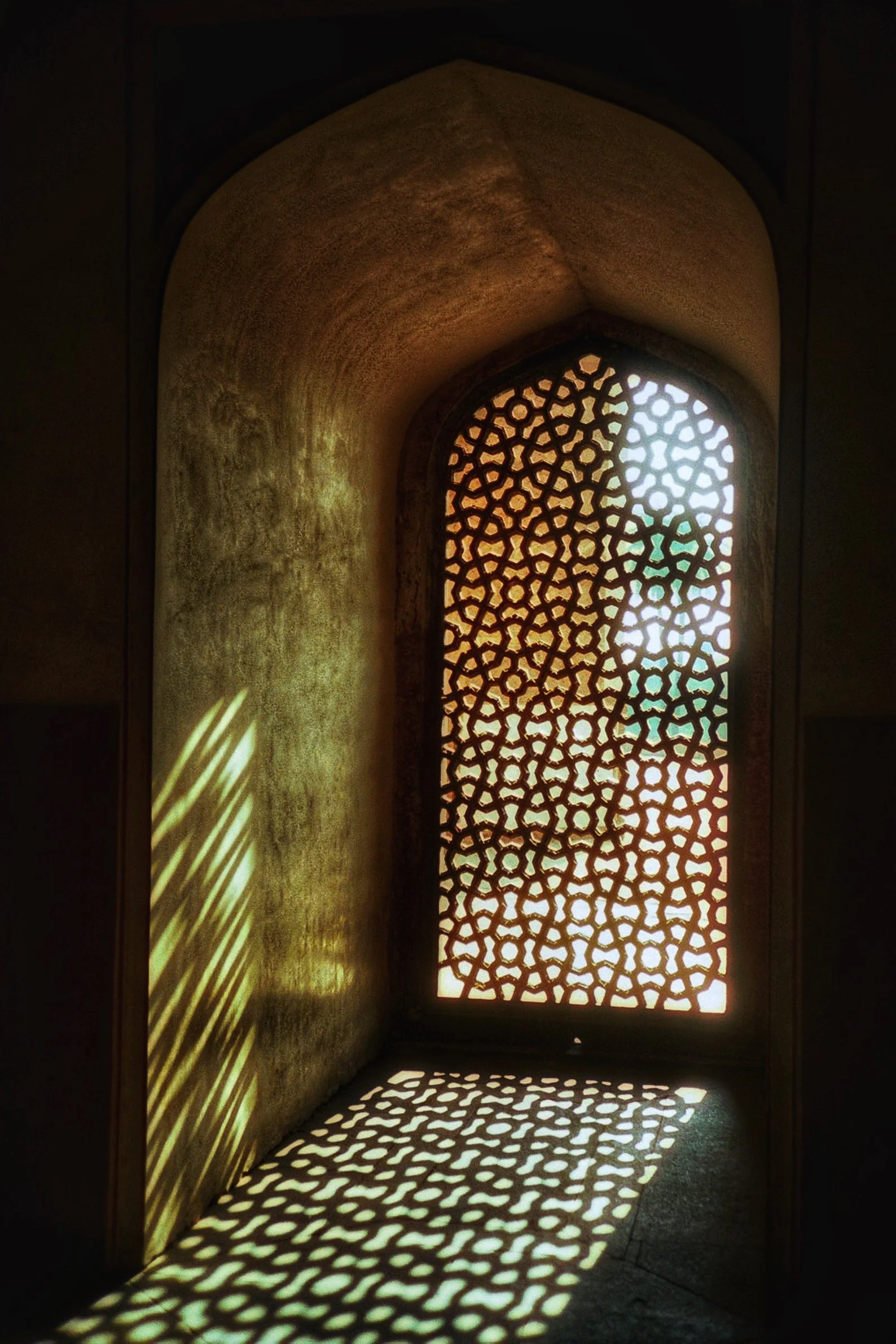 Sunlight passes through an ornate, geometric-patterned screen, casting intricate shadow patterns on the wall and floor inside a dimly lit room.