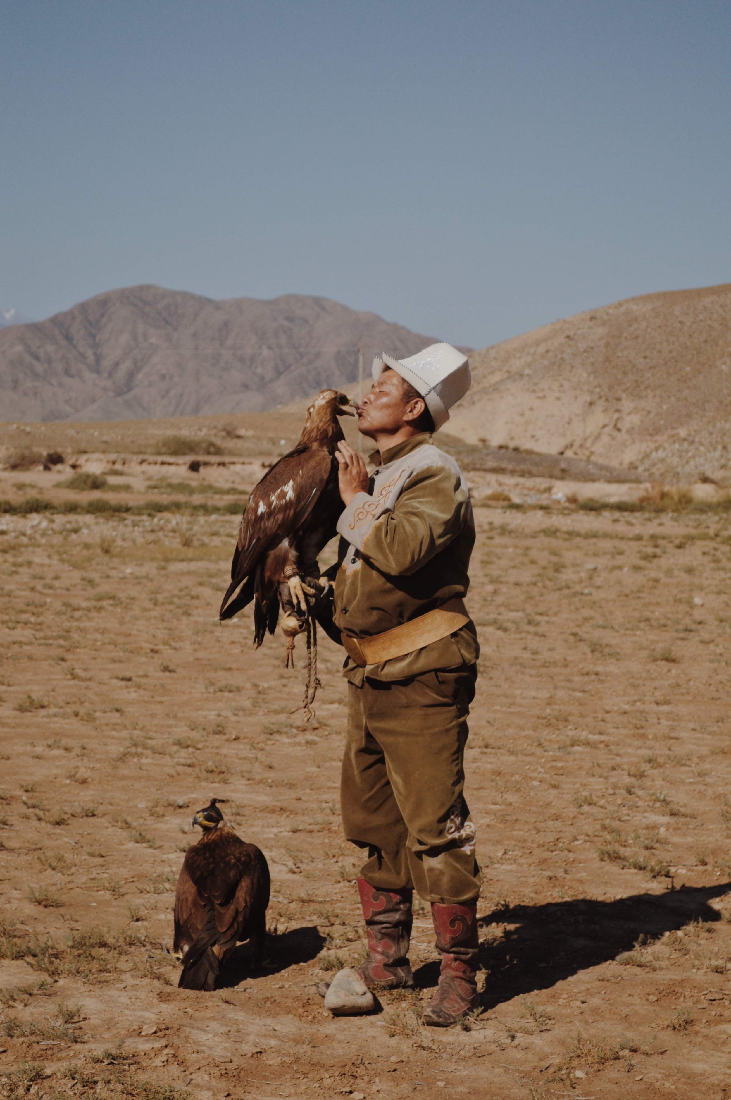 A man in traditional clothing, wearing a large hat, stands in a barren, mountainous landscape holding a bird of prey on his arm. Another bird stands on the ground nearby.