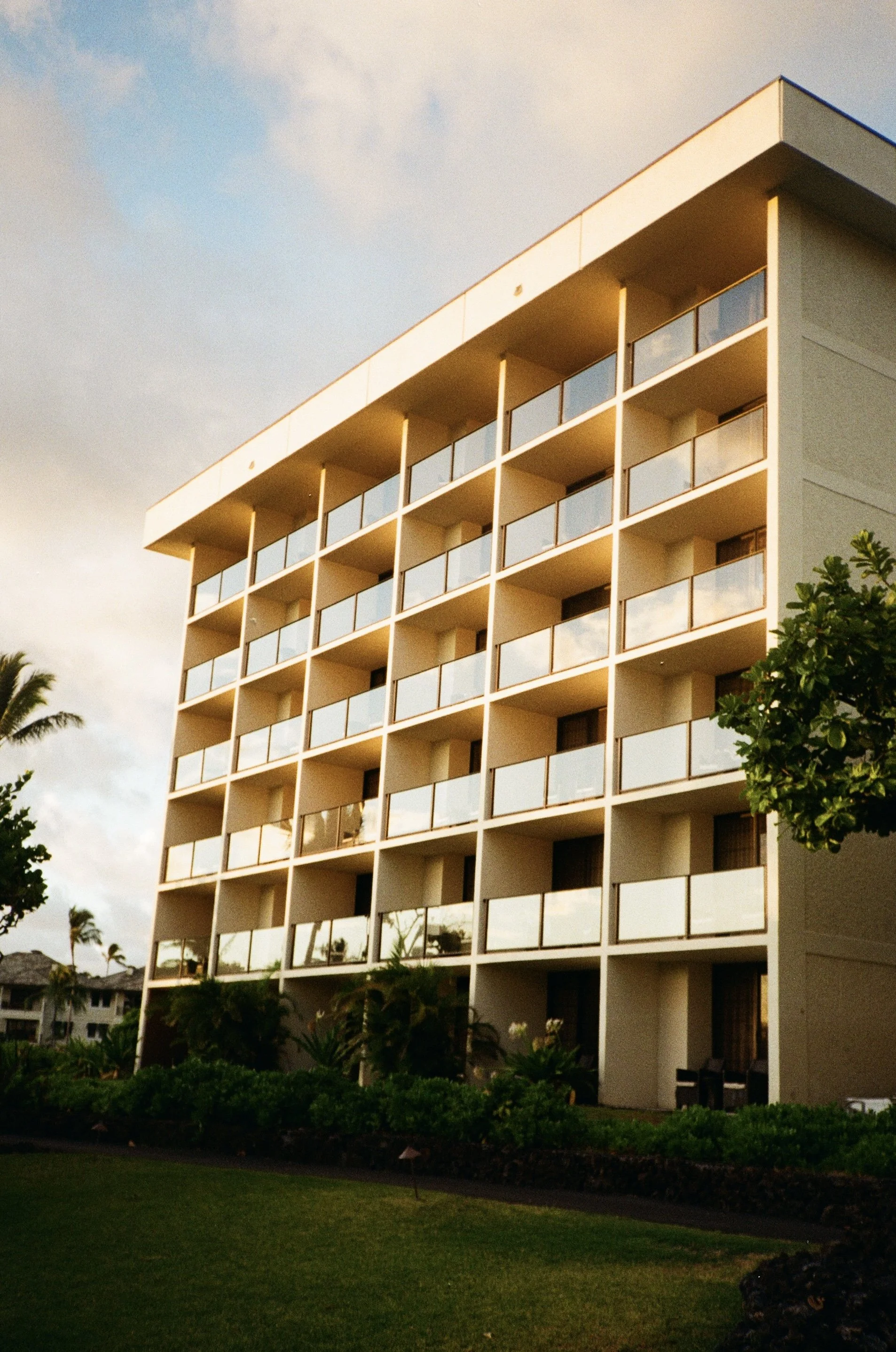 A modern multi-story apartment building with balconies, surrounded by trees and greenery, under a partly cloudy sky in the late afternoon or evening.