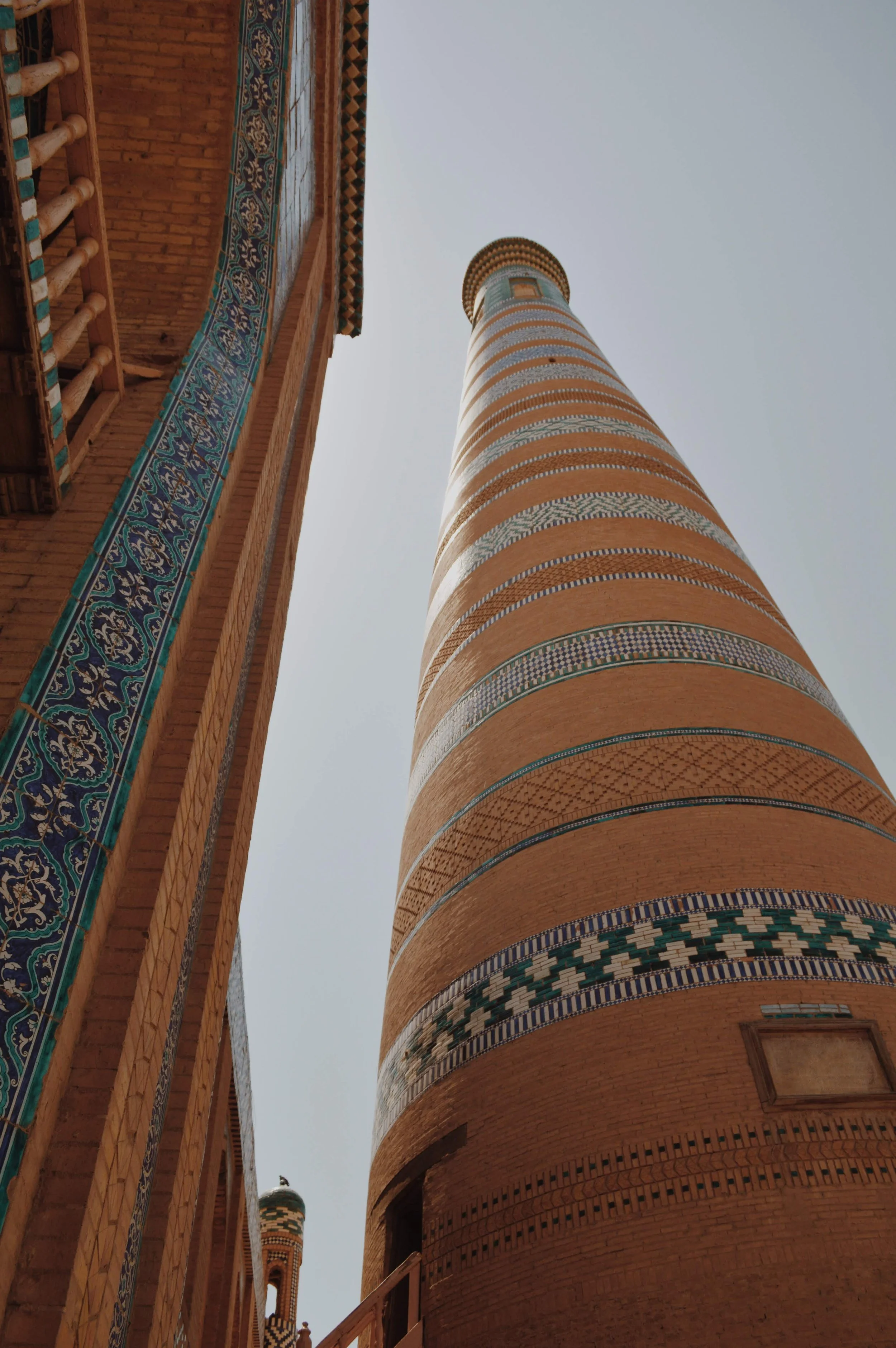 View of a tall, decorative brick tower with alternating patterned bands and a building with ornate tile work and brick walls seen from below.