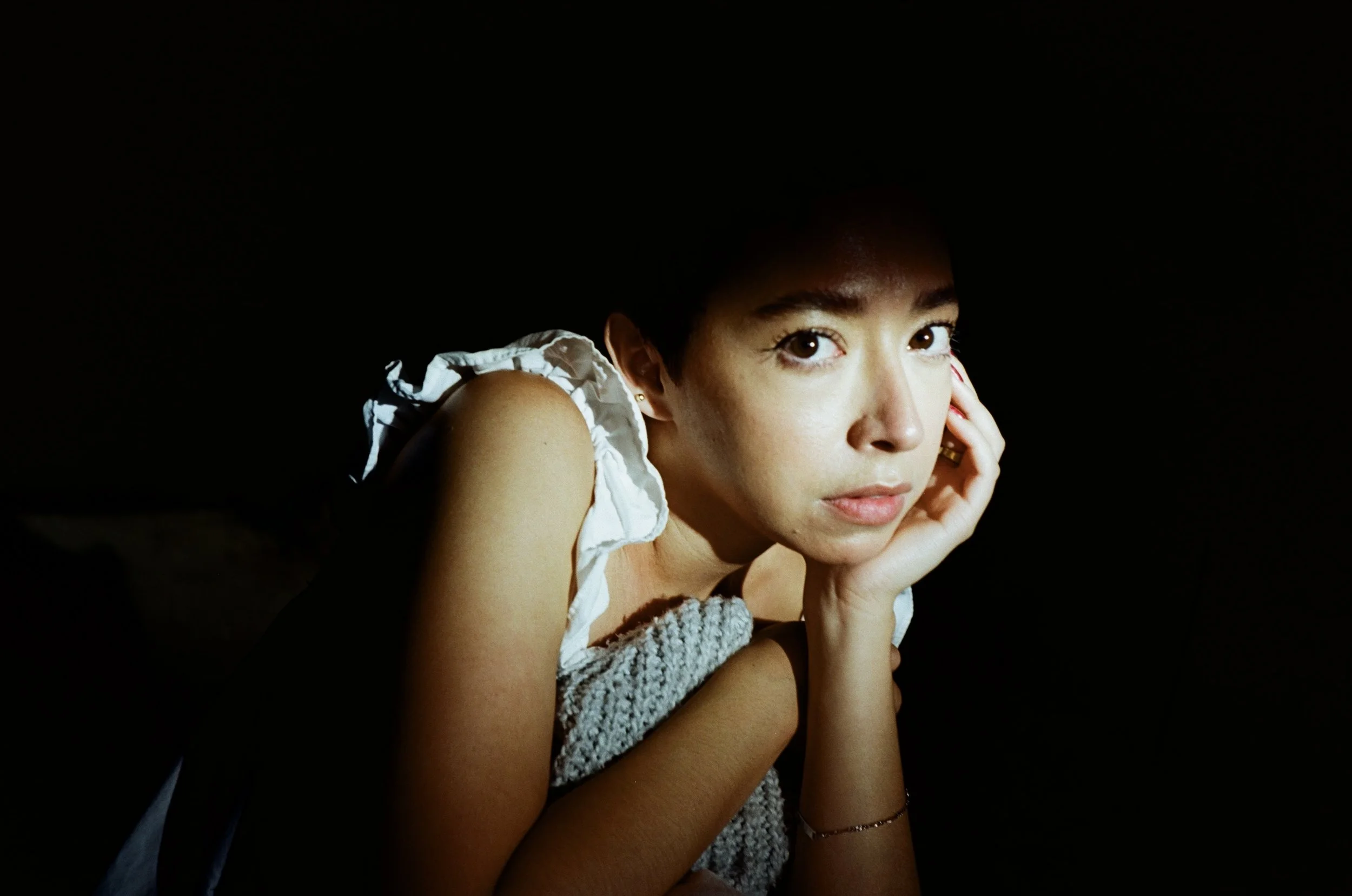 A young woman with brown eyes and light skin, resting her face against her hand, looking into the camera, wearing a sleeveless top with ruffled straps, and a gray knitted sleeve on her arm, against a black background.