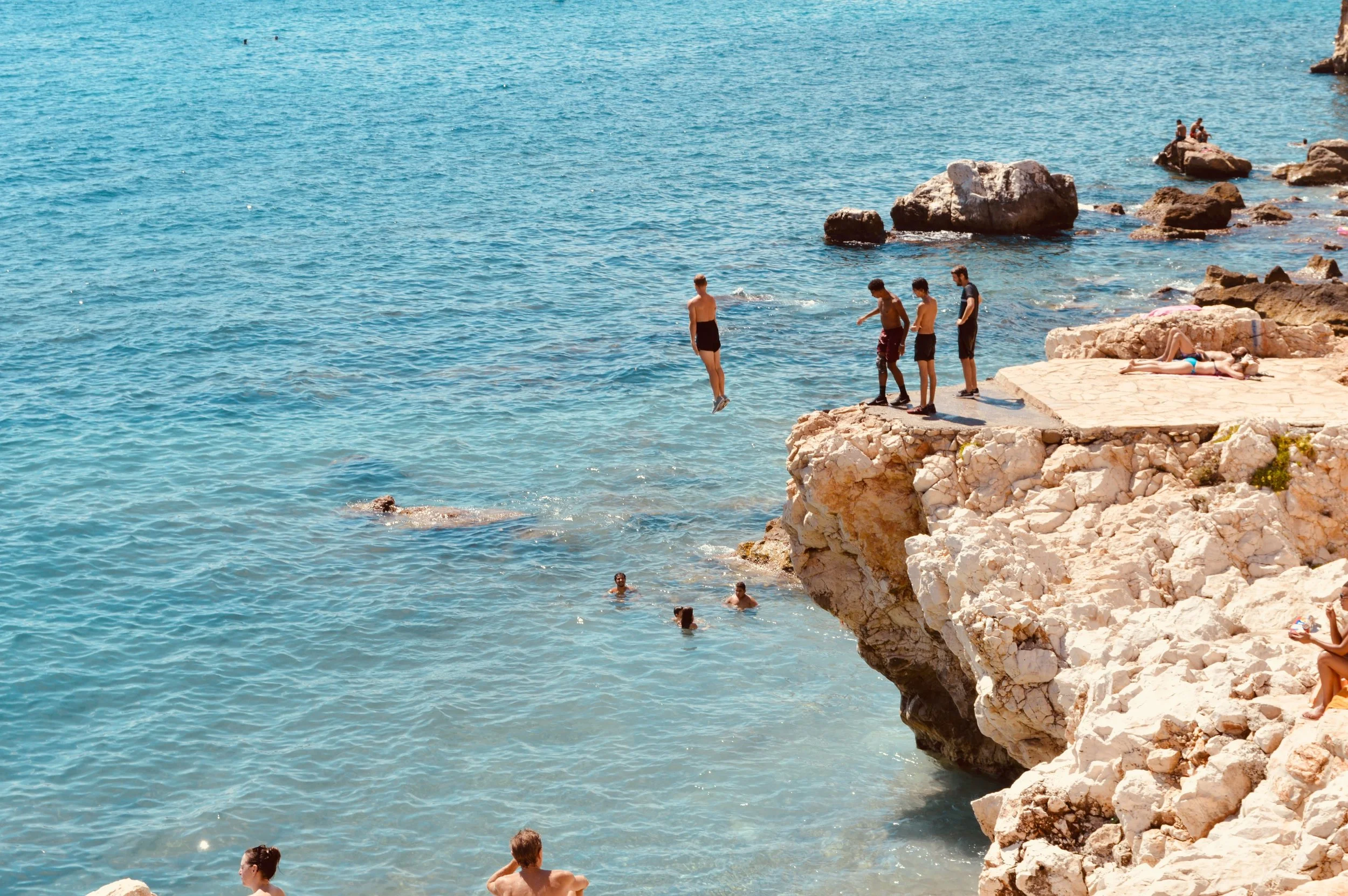 People swimming and relaxing on a rocky coastline with clear blue water and large rocks.