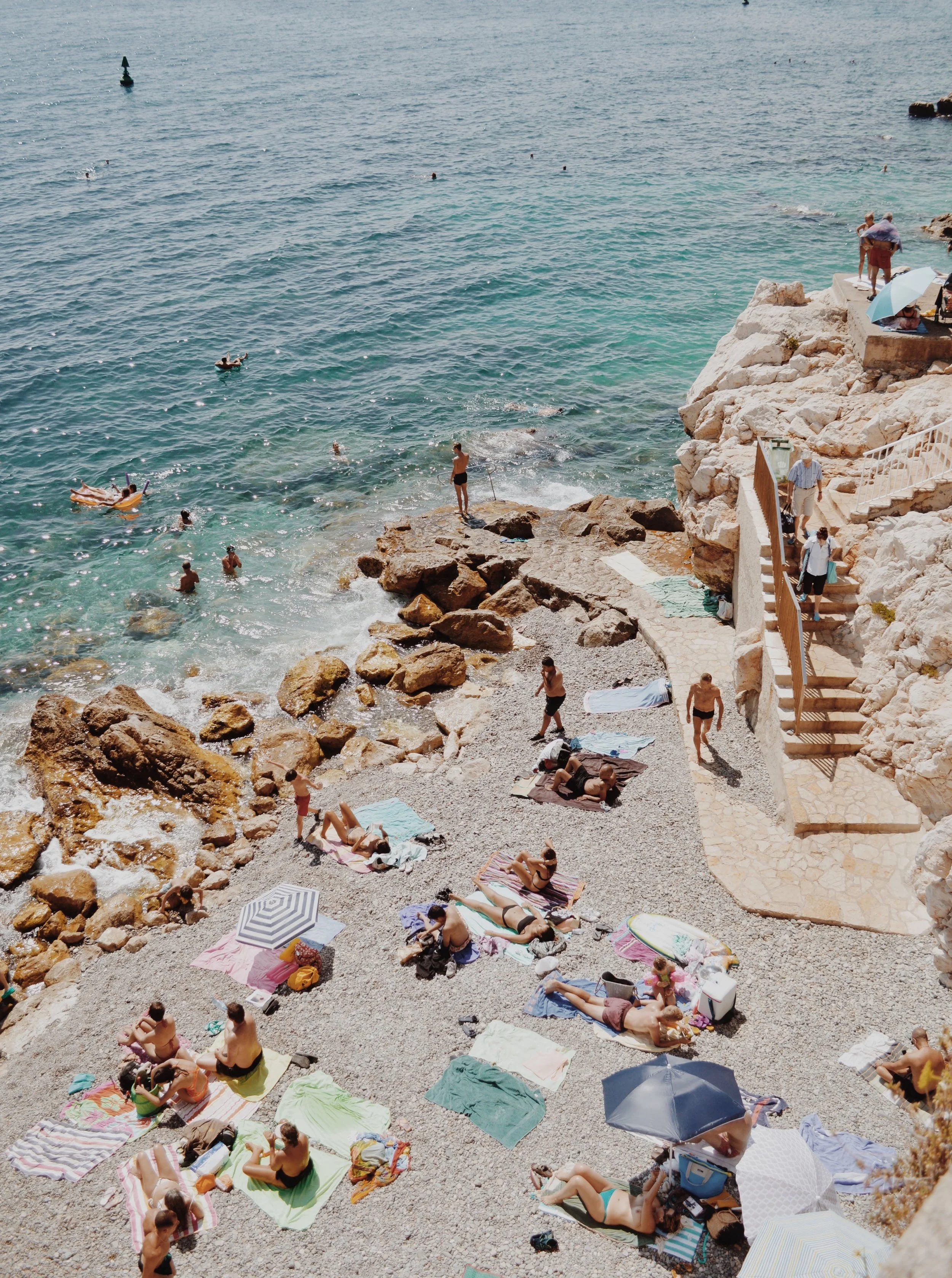 People relaxing on a rocky beach with towels and umbrellas, some swimming and others sitting or walking near the water.