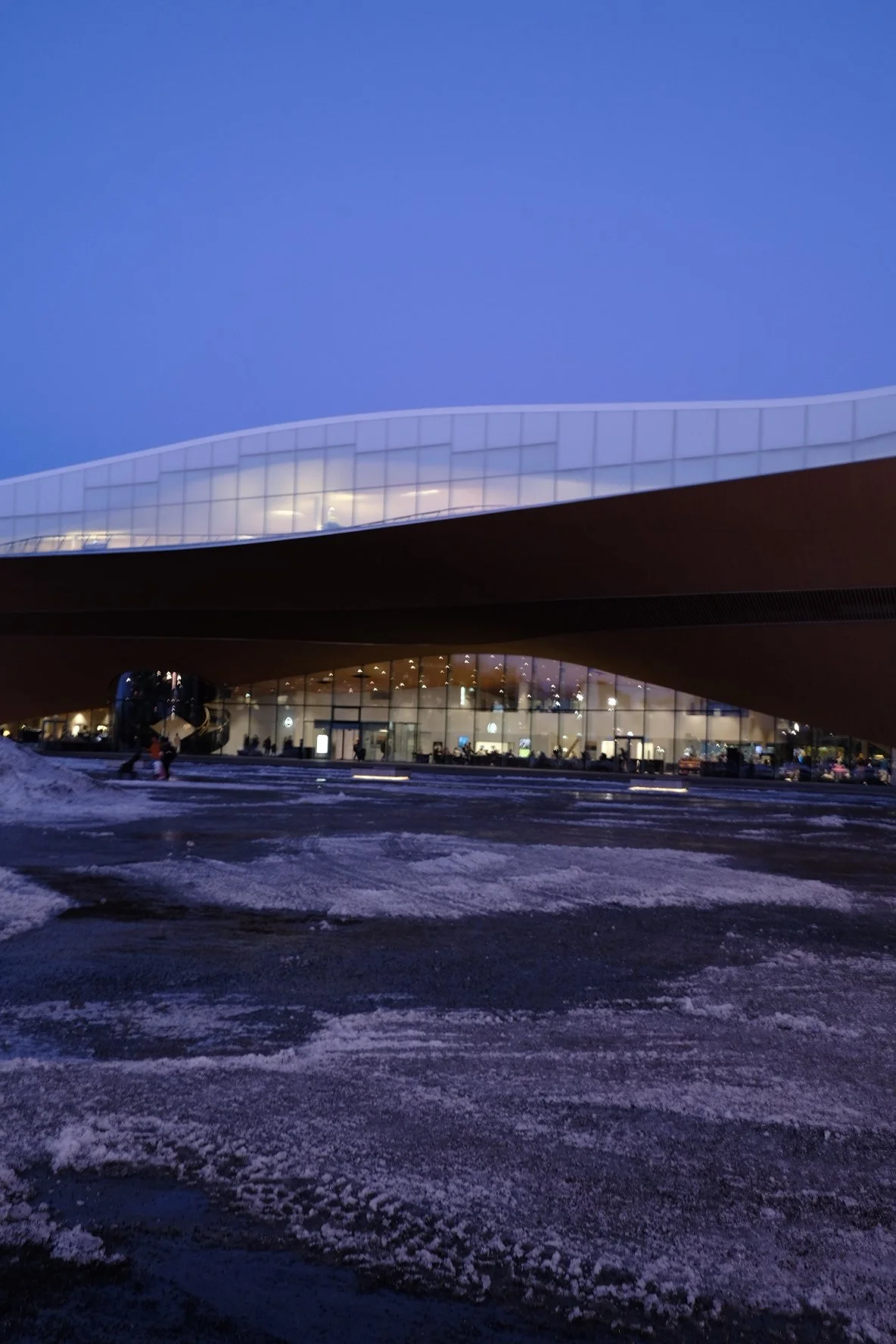 Modern building with large glass windows illuminated from inside, winter conditions with snow and ice on the ground, dark evening or early night sky.