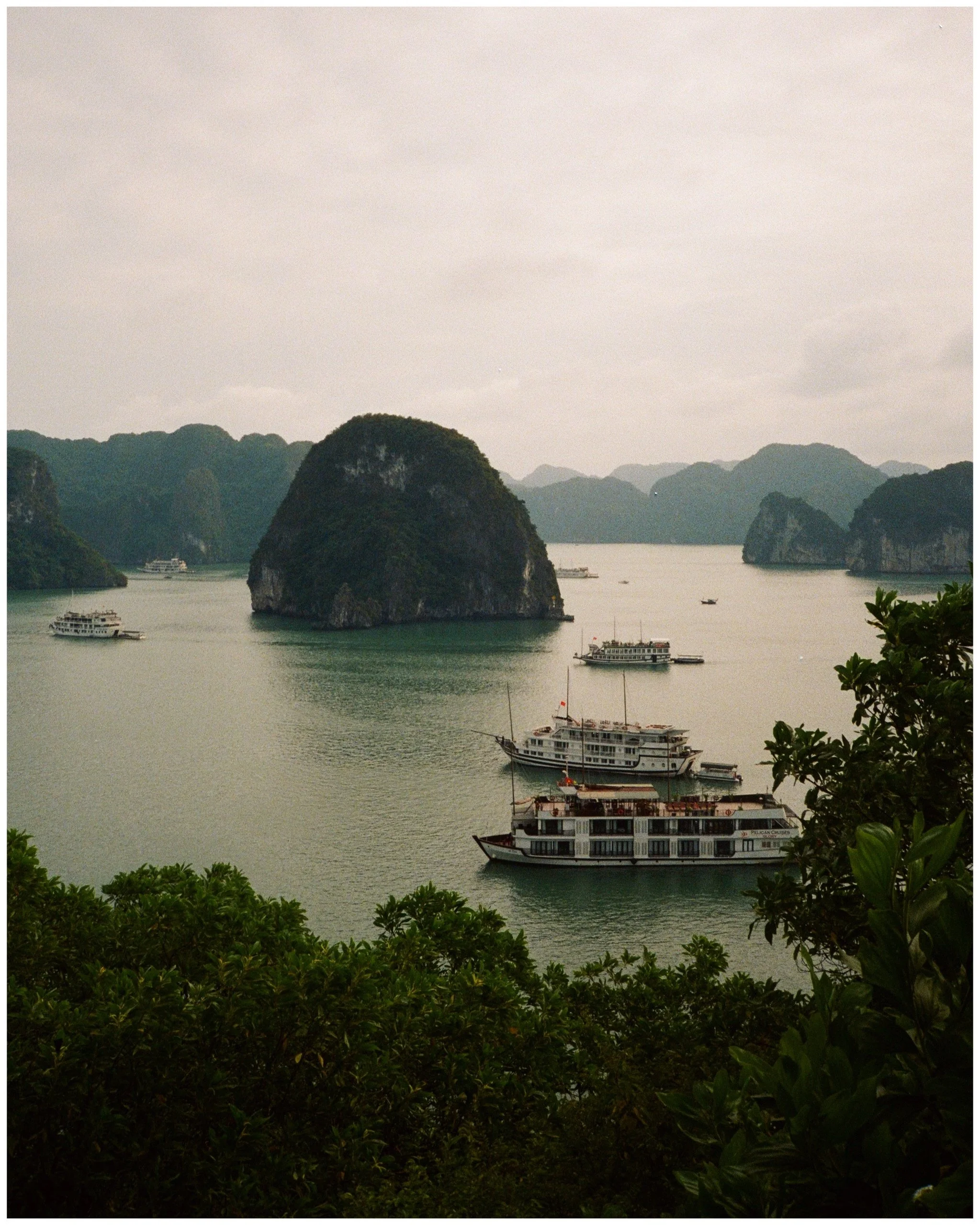 Scenic view of a river surrounded by lush, green, limestone karst mountains with several boats, including a prominent large white cruise ship, sailing on the water at sunset or overcast weather.