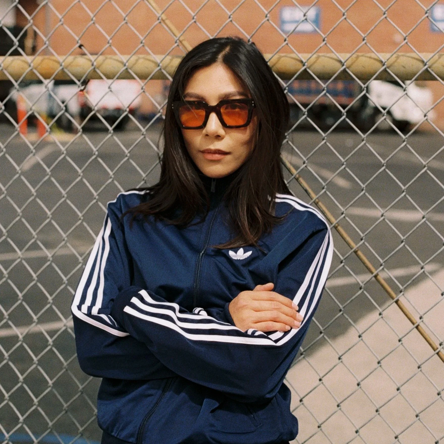 A woman with dark hair wearing large, rectangular sunglasses and a navy blue Adidas jacket with white stripes, standing with her arms crossed in front of a chain-link fence.