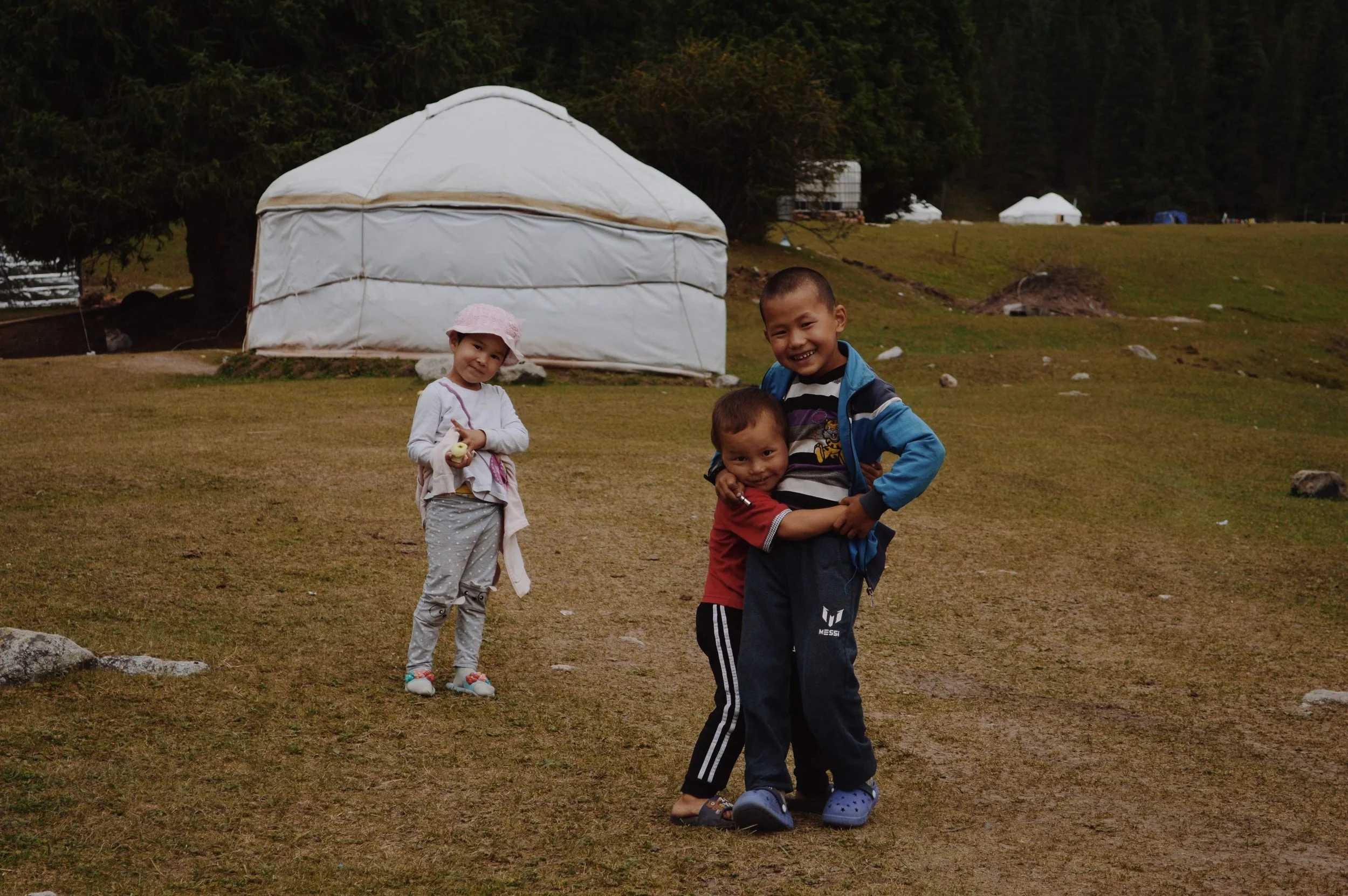 Three children playing outdoors near a yurt, with one child on a scooter, one girl holding a toy, and another smiling child hugging a smaller child, surrounded by grass and trees in a rural setting.