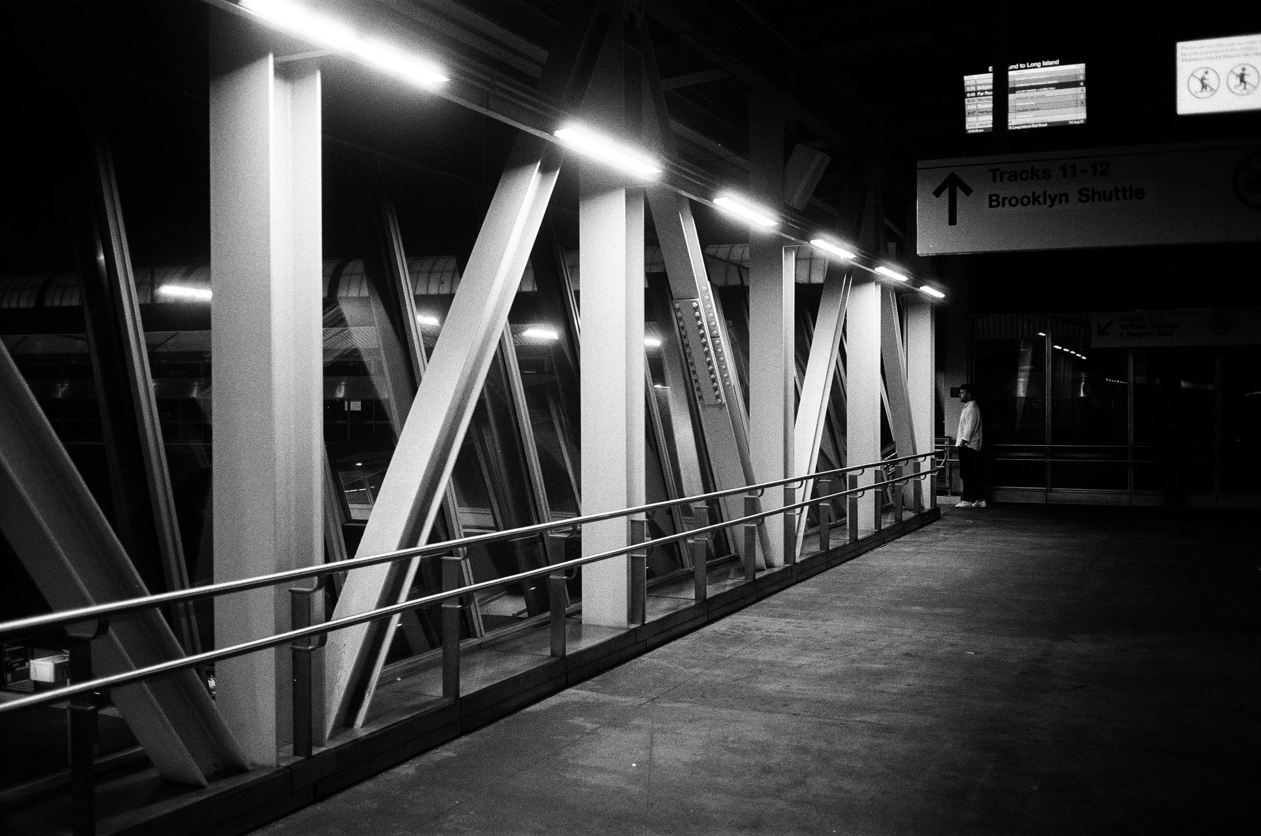 A black and white photo of an airport terminal, showing a row of large, angled windows with structural supports. There is an empty walkway with a railing on the right, and a person standing in the distance near the windows. Overhead signs indicate tr