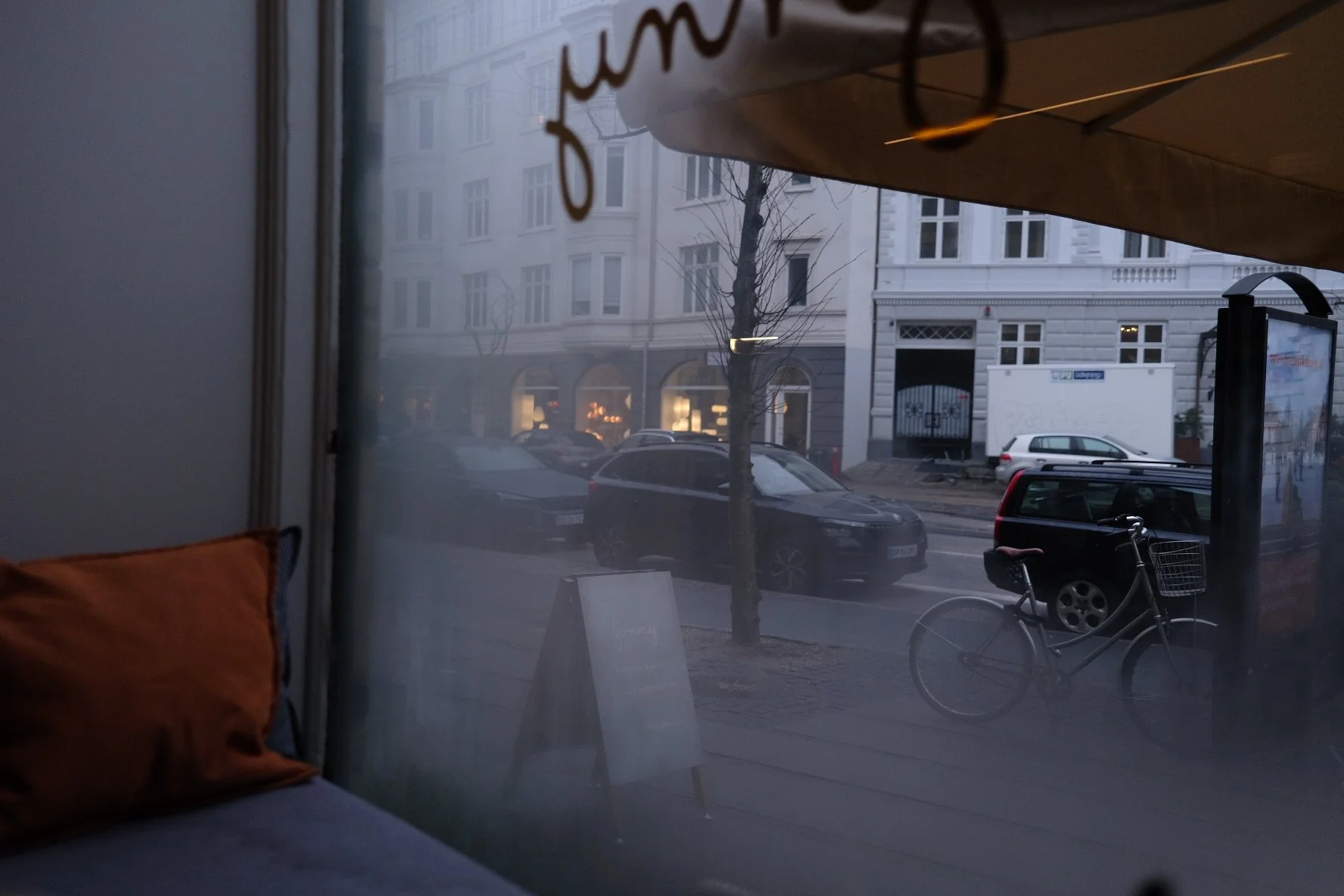 View of a city street through a window, with parked cars, a bicycle, trees, and a building with white facade and arched windows across the street.